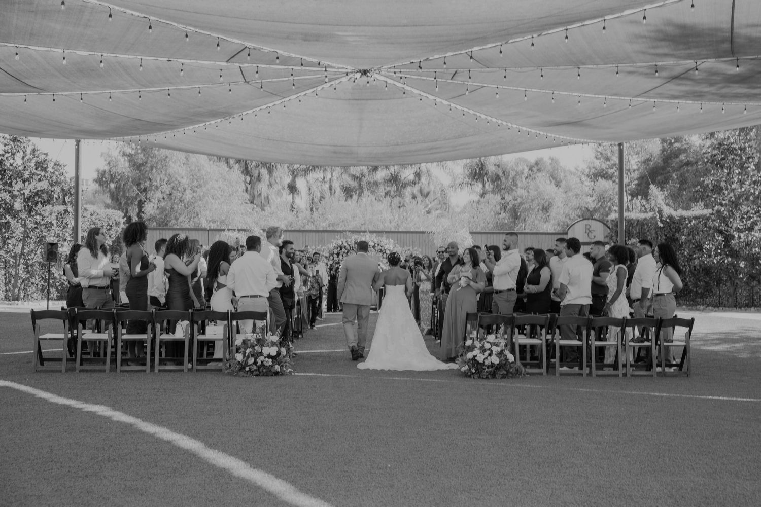 Black and white outdoor wedding ceremony under a canopy with string lights, guests seated in rows facing the couple at the altar.