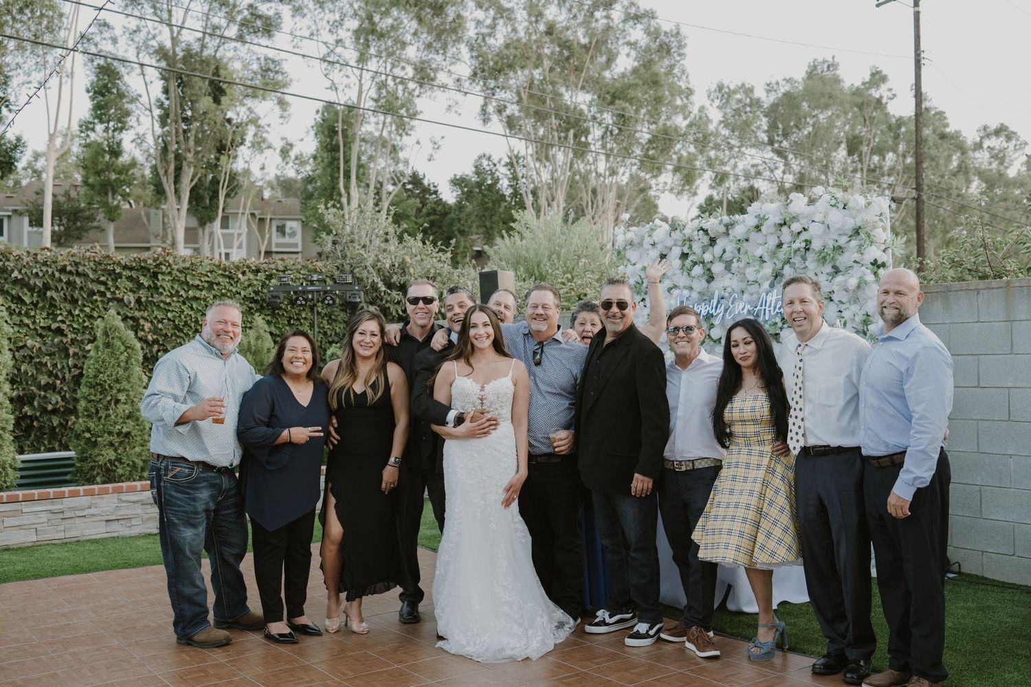 A group of wedding guests posing with the bride in a white gown at an outdoor garden wedding venue.