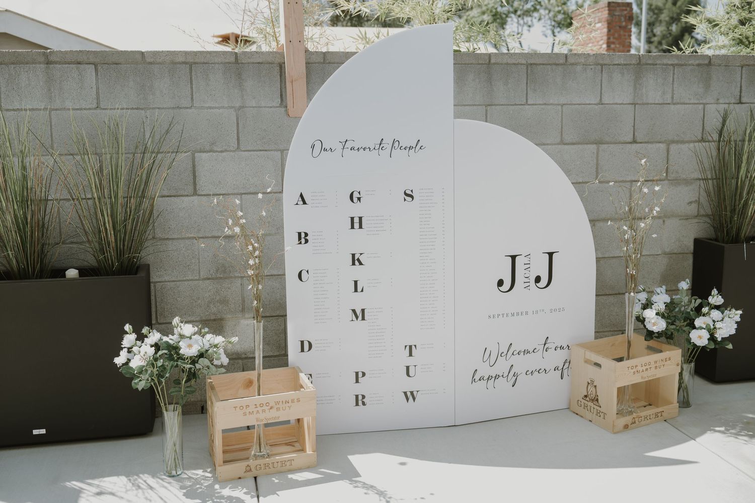 Elegant wedding seating chart display with monogram J&J on white arch panels, flanked by wooden crates and floral arrangements.