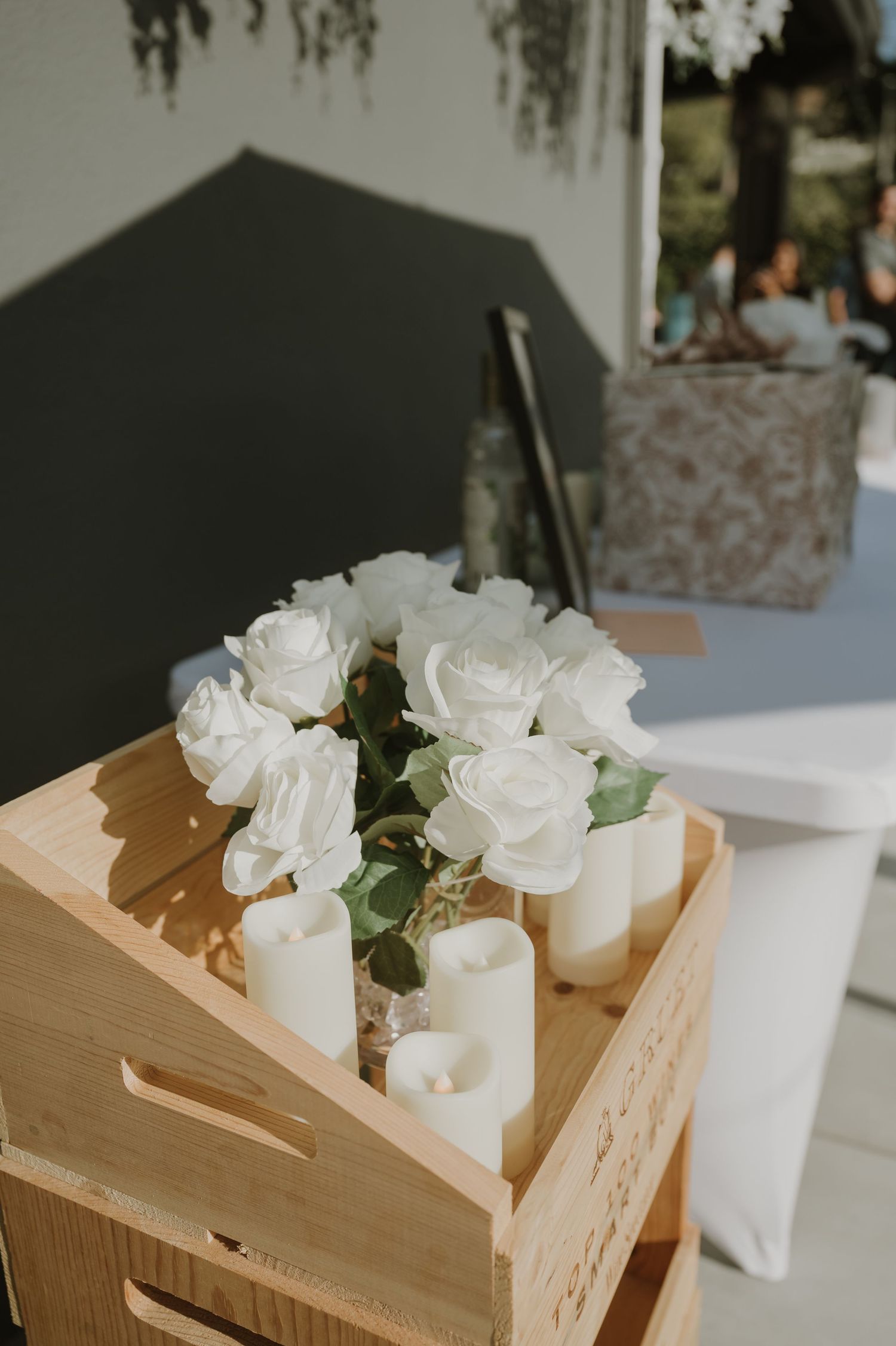 White roses and pillar candles arranged on a rustic wooden crate at an outdoor wedding reception.