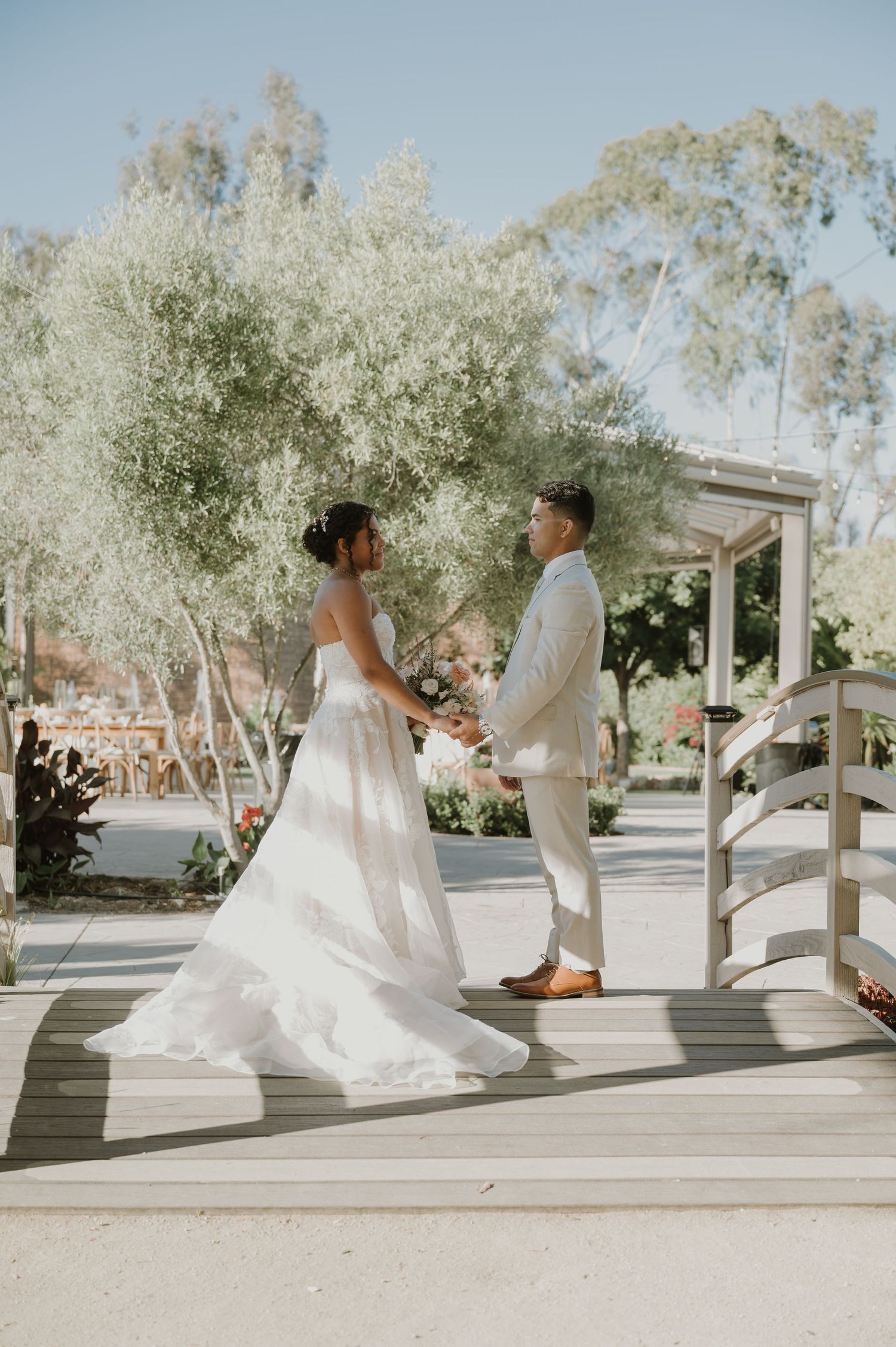 Bride and groom holding hands on a wooden bridge during outdoor wedding ceremony among olive trees.