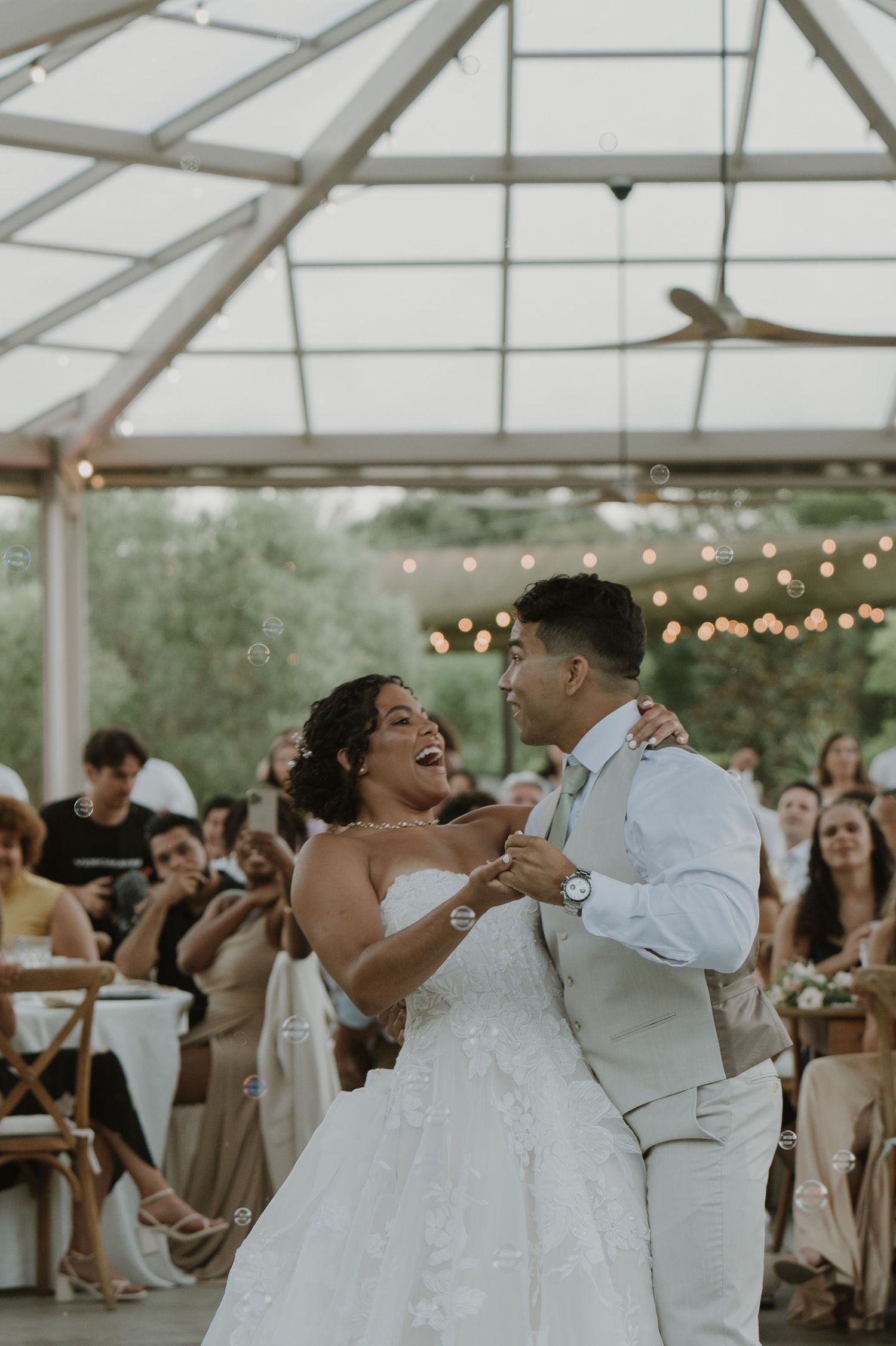 Bride and groom share joyful first dance under glass ceiling with string lights and bubbles.
