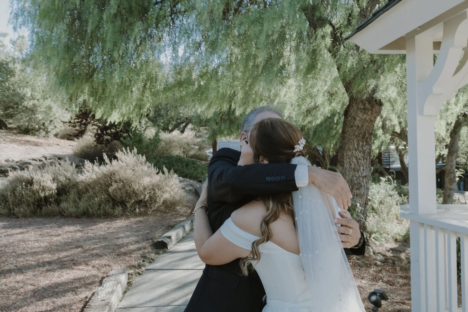 Bride and groom share a tender embrace outdoors near a white gazebo surrounded by lush green trees on their wedding day.