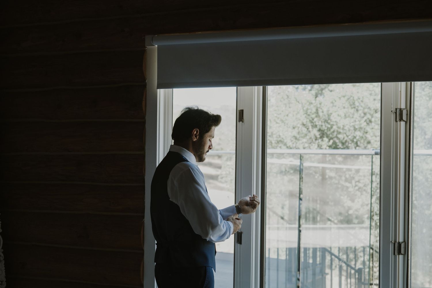 Groom in vest gazing out window holding cufflinks while preparing for wedding ceremony.