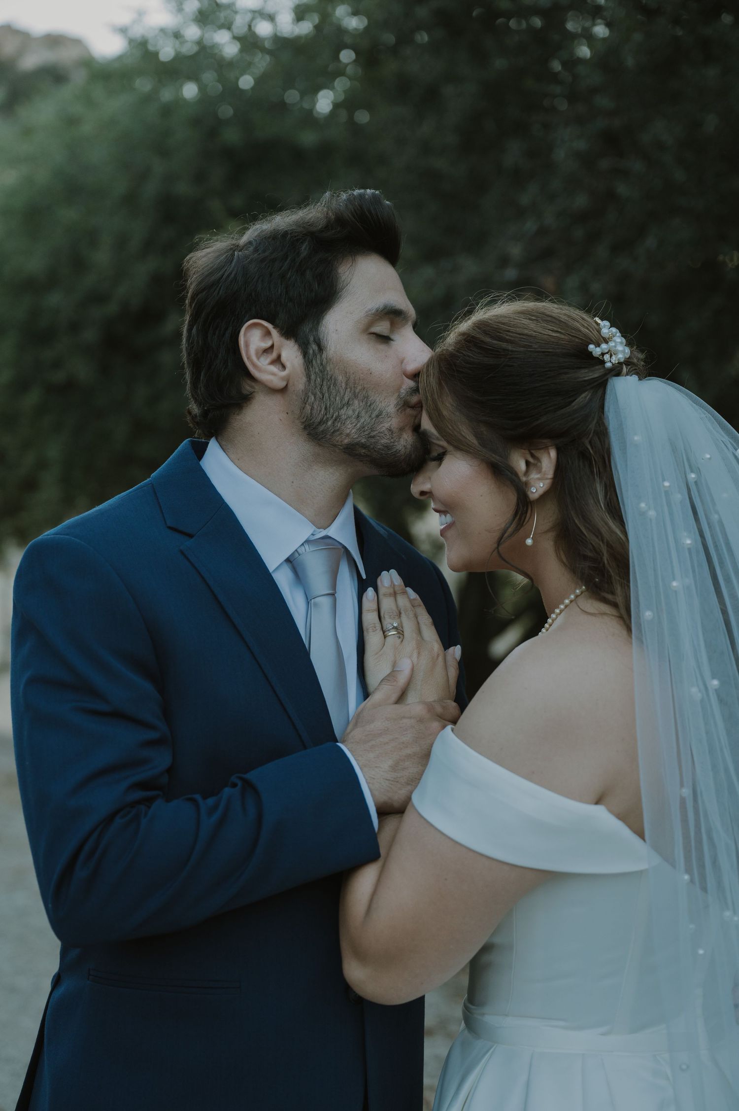 Groom in navy suit kisses bride on forehead as she smiles, hands clasped, wearing off-shoulder gown and veil.