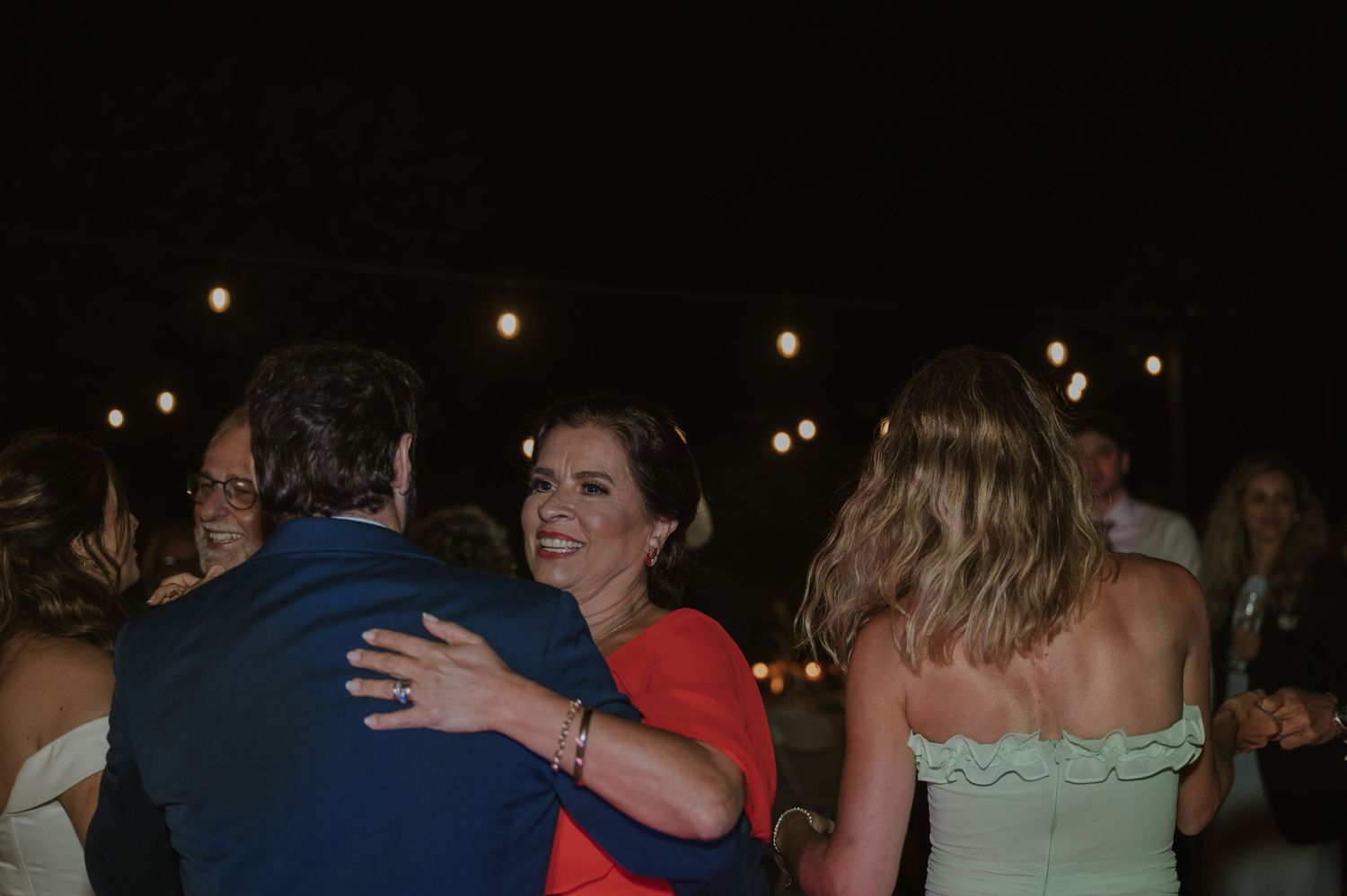 Guests dancing and embracing at an outdoor wedding reception with string lights glowing in the night sky.