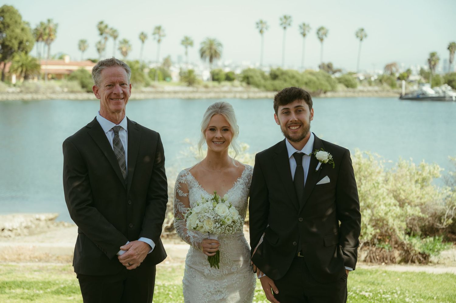 Bride in lace gown with groom and officiant posing outdoors near waterfront with palm trees at wedding ceremony.