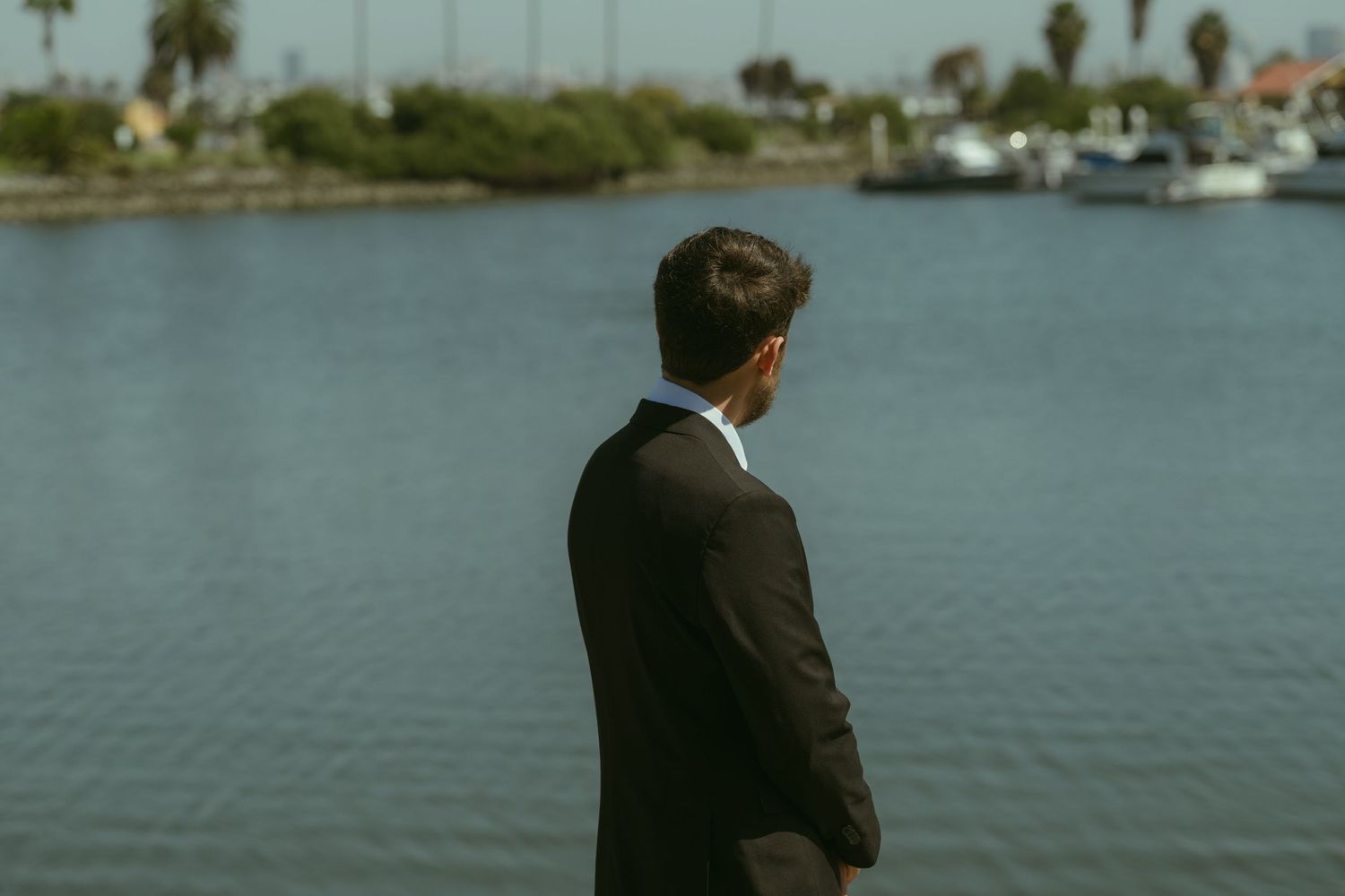 Groom in dark suit standing with back to camera overlooking marina waterfront.