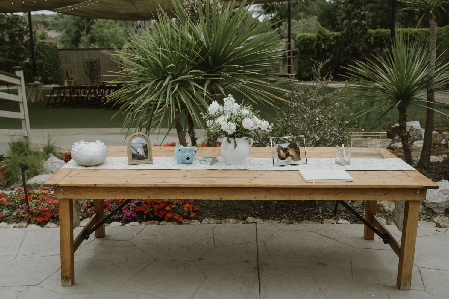 Rustic wooden farm table outdoor wedding display with white flowers, photo frames, and decorative items on stone patio.