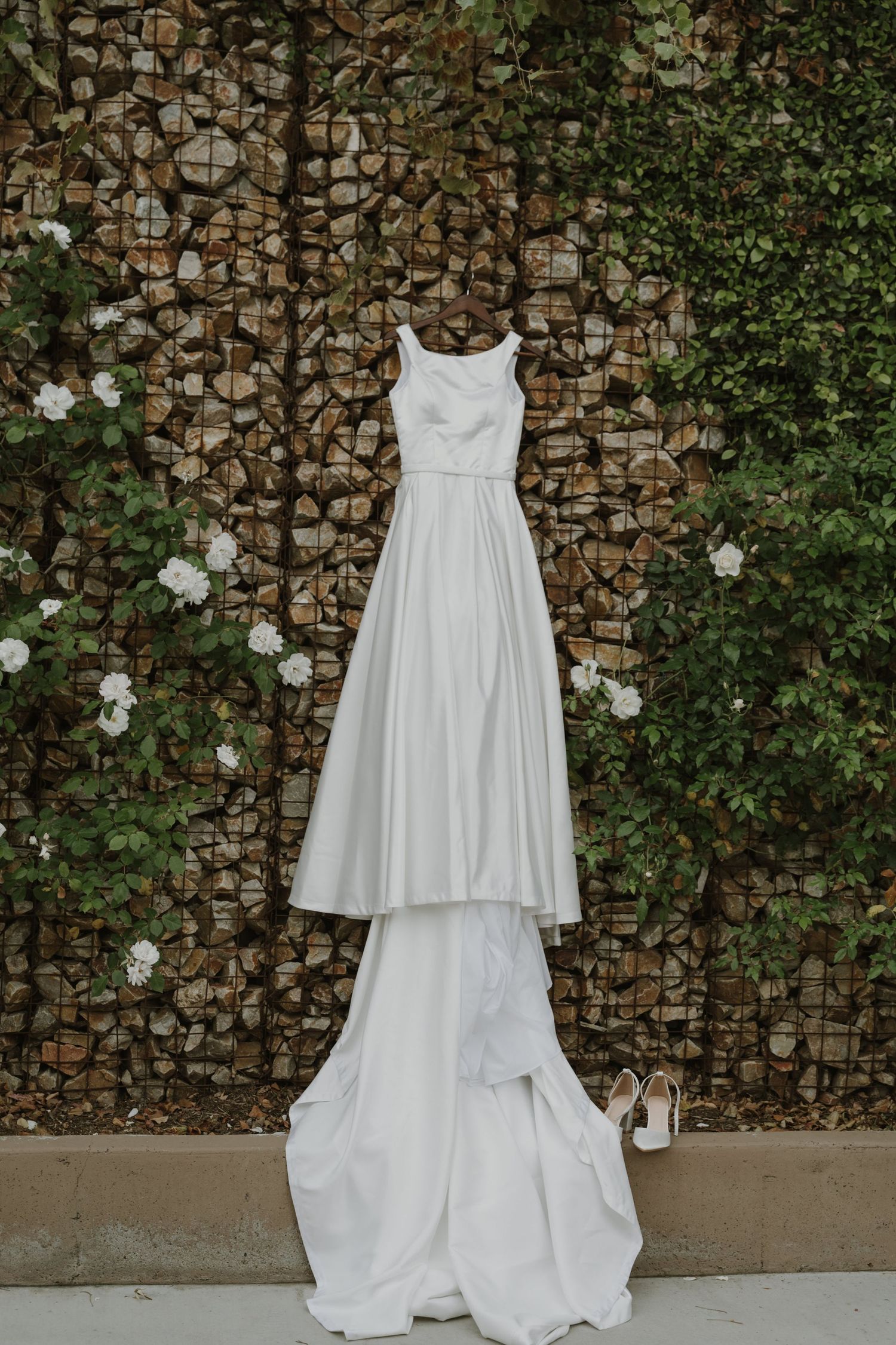 Elegant white wedding gown hanging outdoors against a stone wall with lush greenery and white flowers.