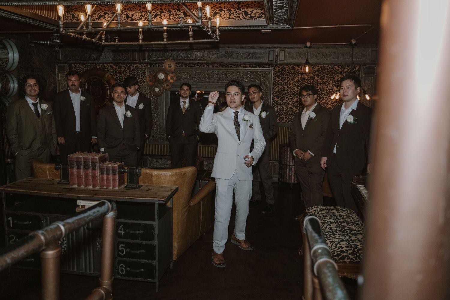 Groom in white suit poses with groomsmen in brown suits inside a rustic, ornate venue with vintage decor.