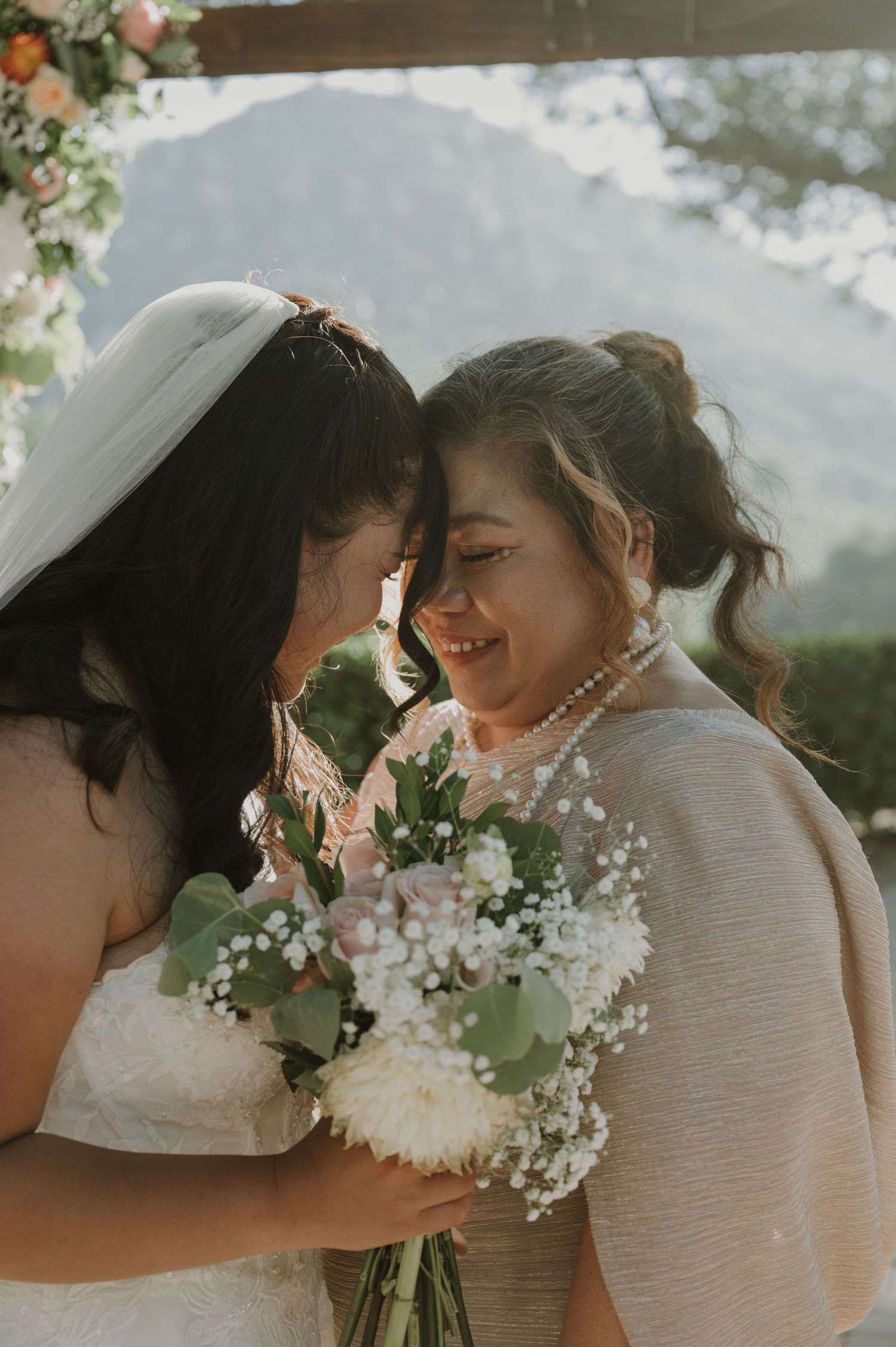 Two brides share an intimate moment touching foreheads, holding a white floral bouquet at an outdoor wedding ceremony.