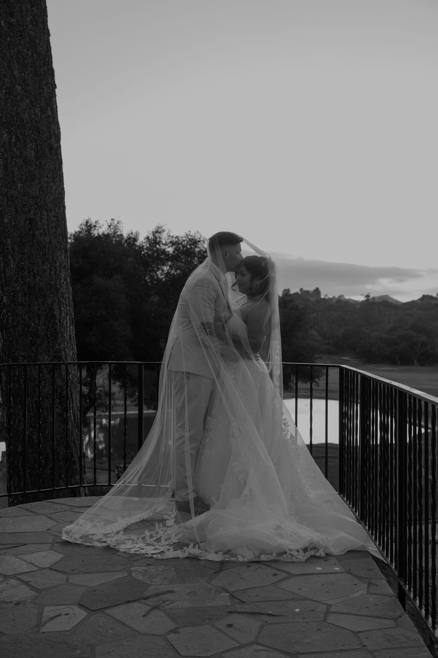 Black and white photo of a bride and groom sharing a kiss on a balcony at dusk, her veil flowing elegantly.