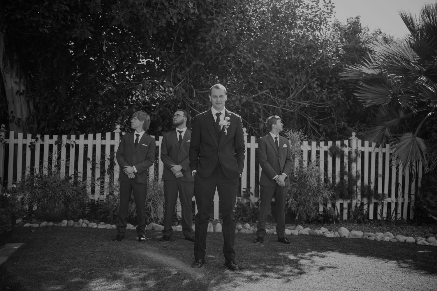 Groom stands with groomsmen in black suits near a white picket fence in a black and white wedding photo.