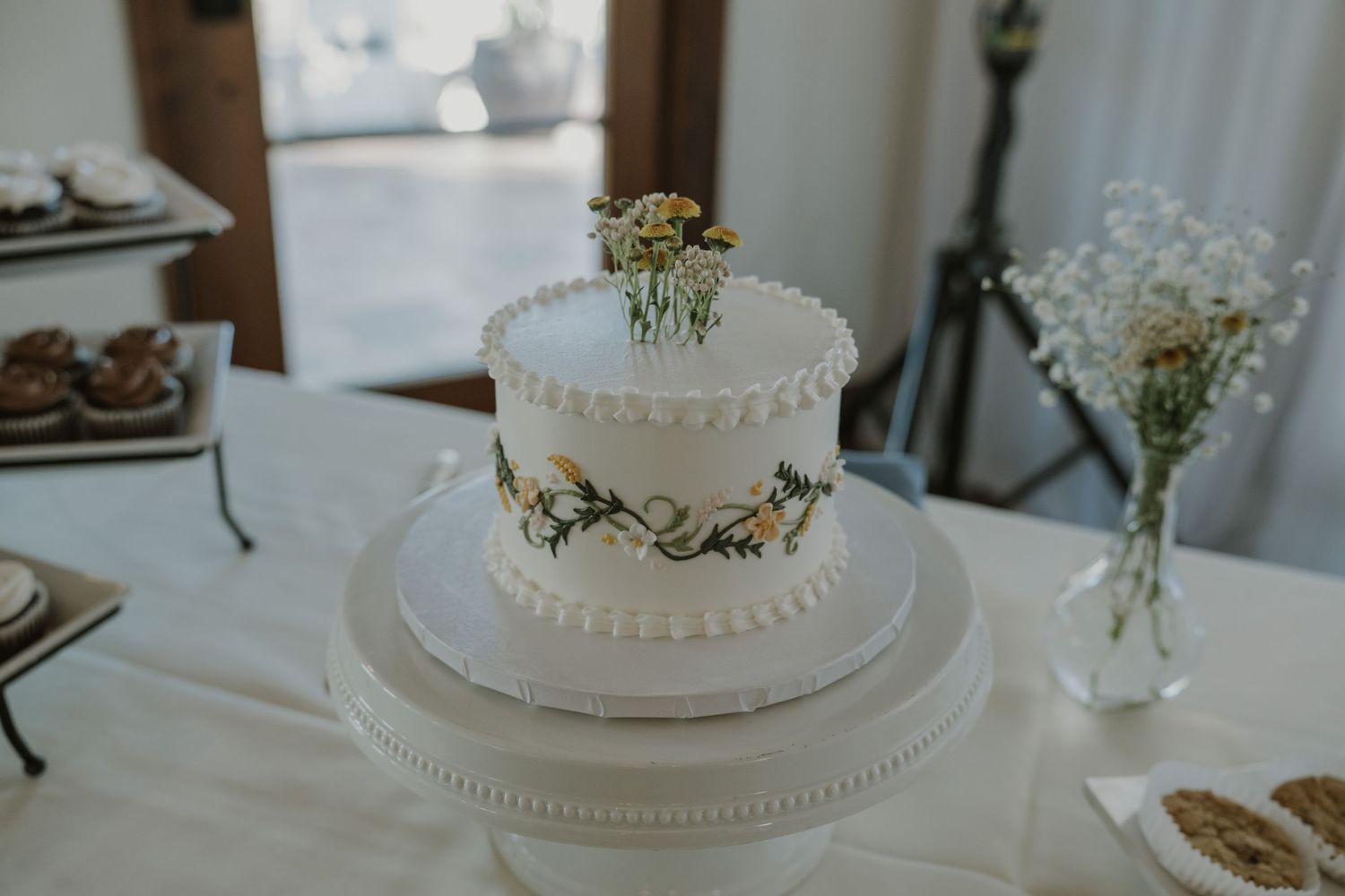 Elegant two-tier white wedding cake with floral topper and yellow flower accents on a white cake stand.