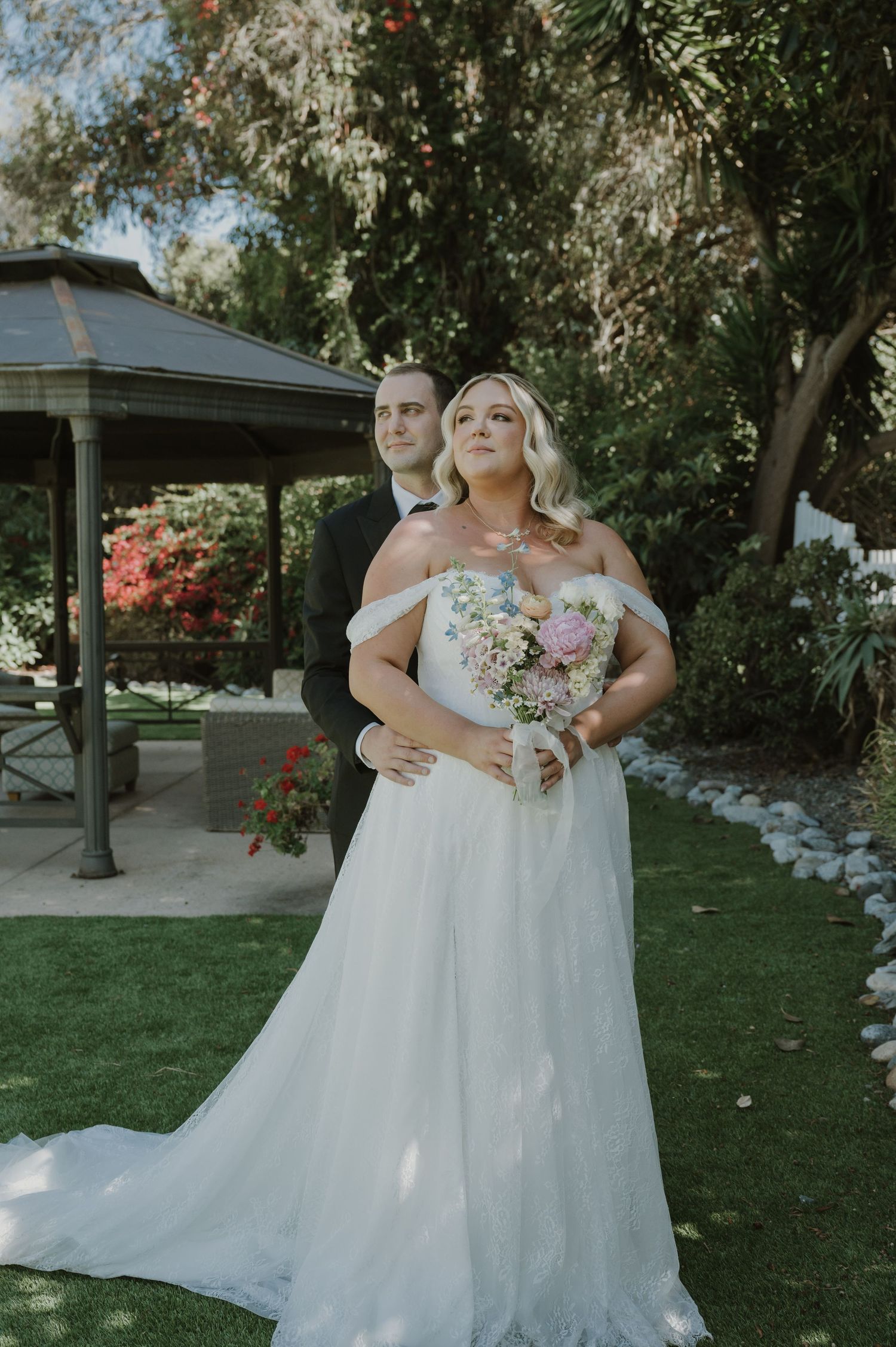 Bride in off-shoulder white gown holding bouquet with groom in black suit, posing in lush garden near gazebo.