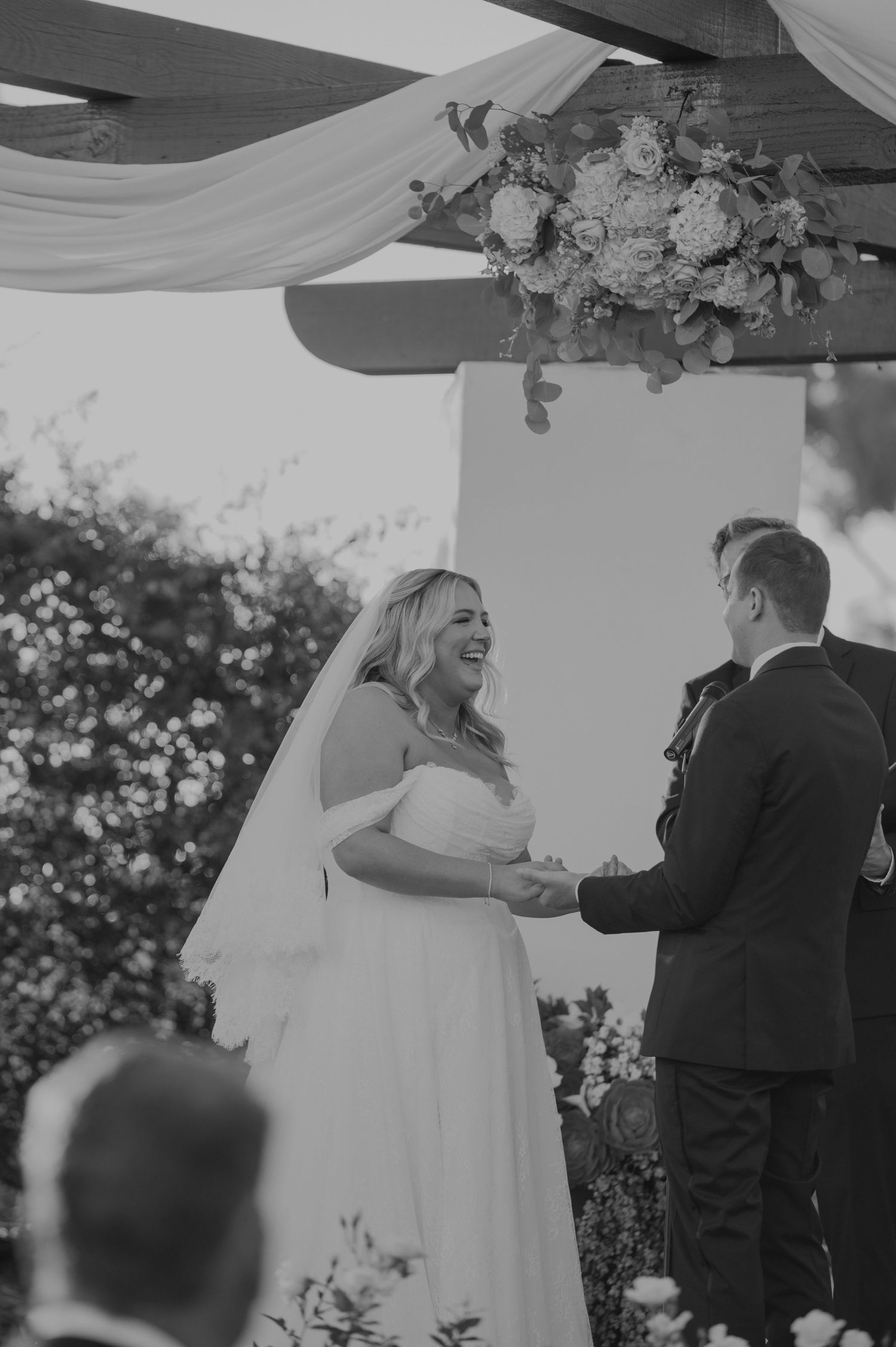 Black and white photo of a laughing bride holding hands with groom at outdoor wedding ceremony under floral arch.
