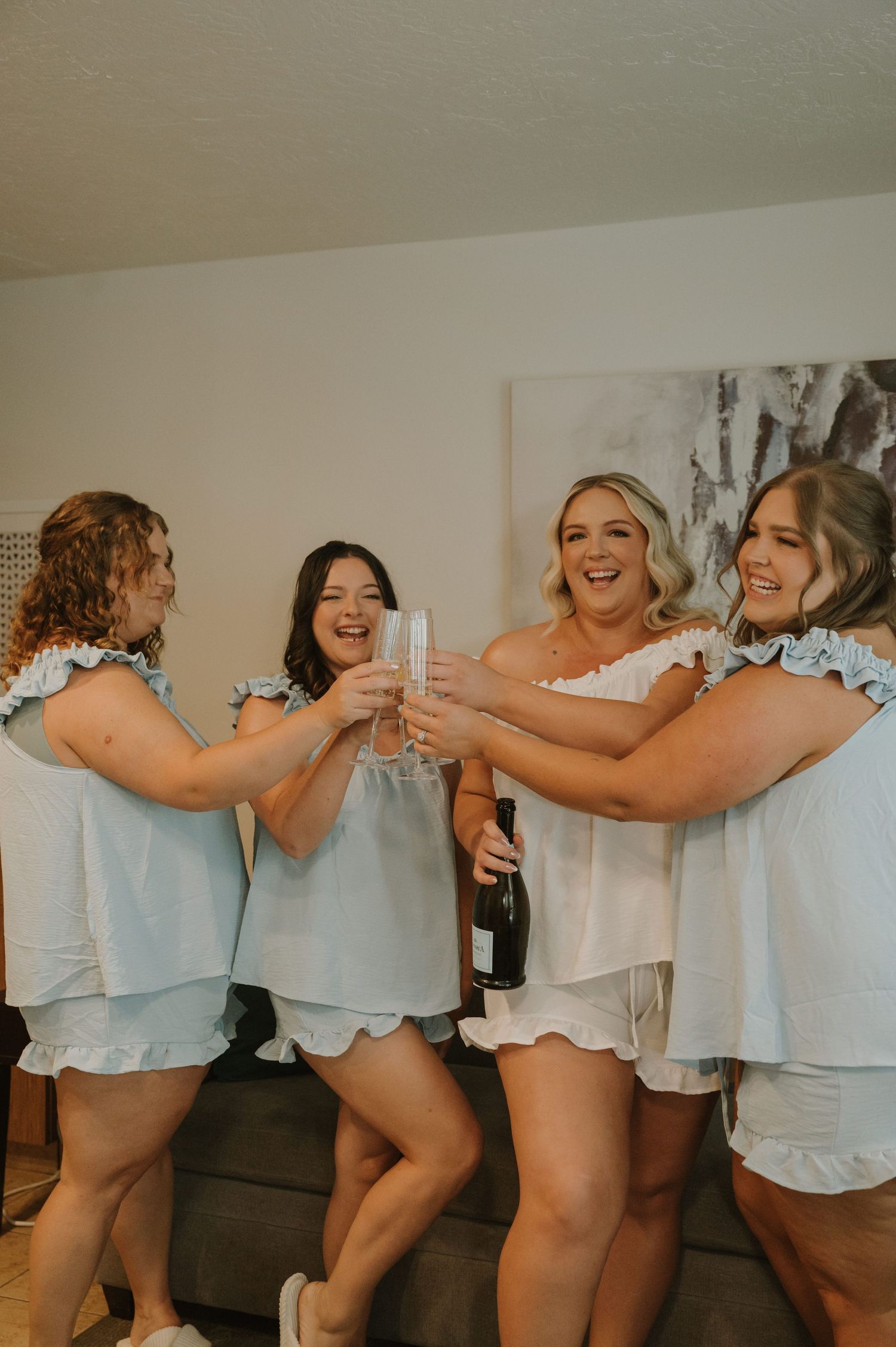 Four bridesmaids in light blue matching sets toasting champagne glasses with a laughing bride in white during wedding preparations.
