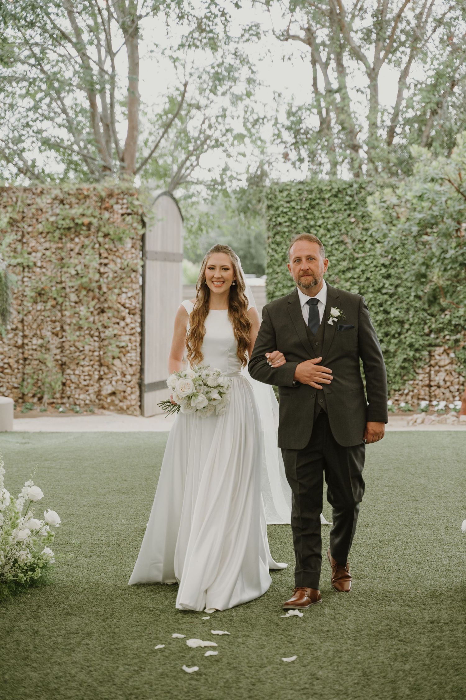 Bride in white gown holds bouquet while walking down aisle with escort at outdoor wedding ceremony.