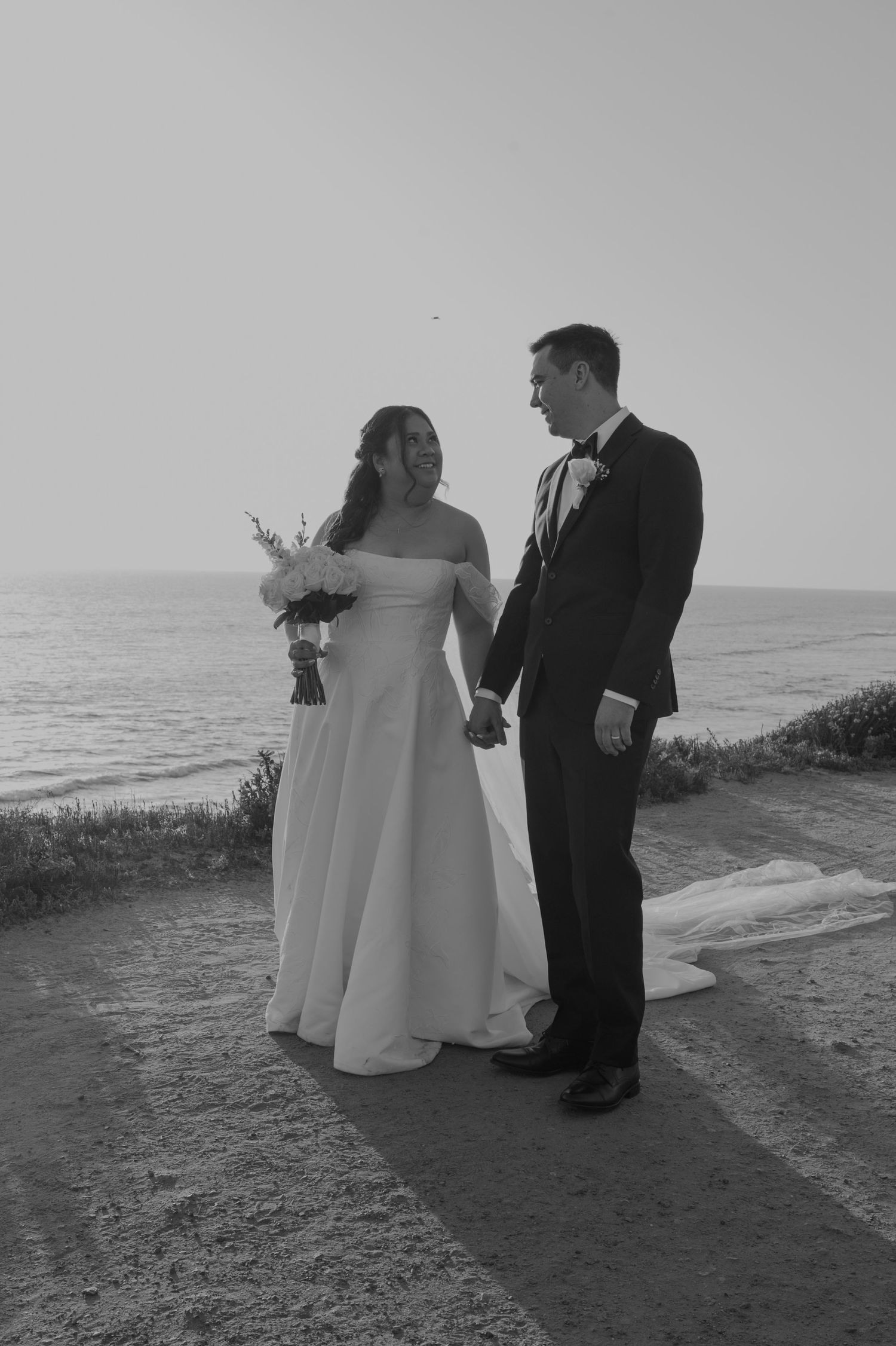 Black and white photo of a bride and groom holding hands by the ocean, bride holding a bouquet in a strapless gown.