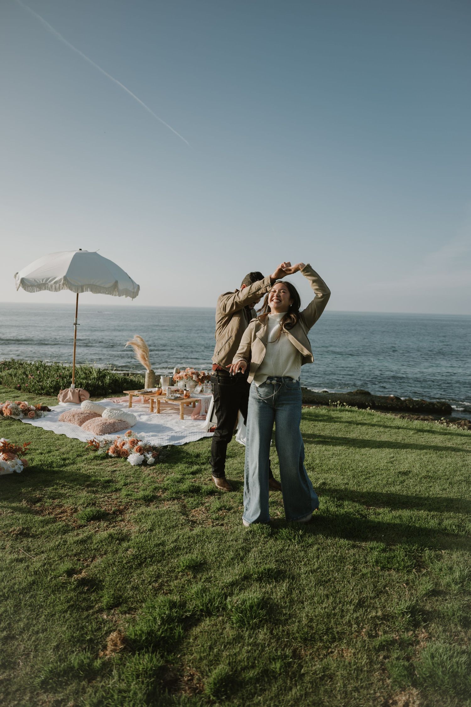 A couple shares a romantic moment on a clifftop overlooking the ocean with picnic setup nearby.