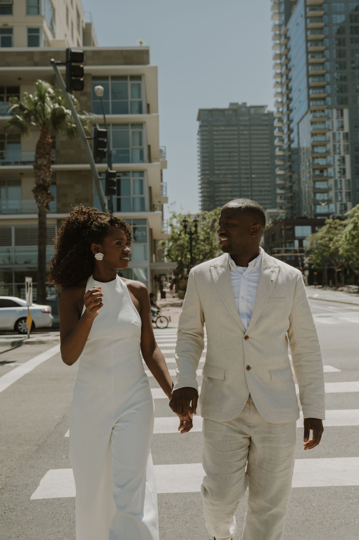 A couple in crisp white attire walking together on a city street with palm trees and buildings.