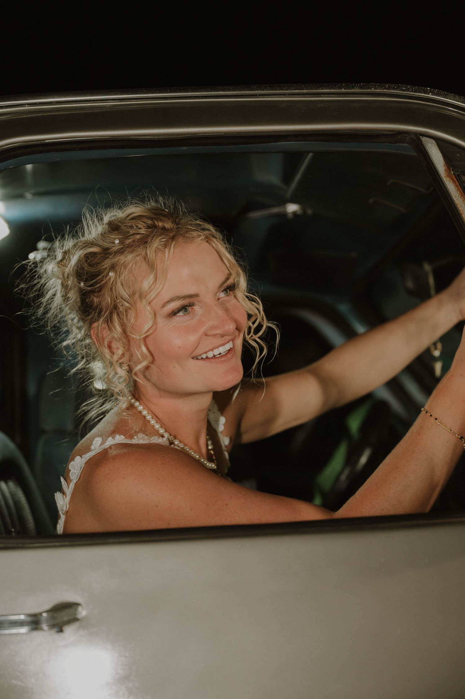Person with curly hair smiles while sitting in a classic car during wedding celebrations.