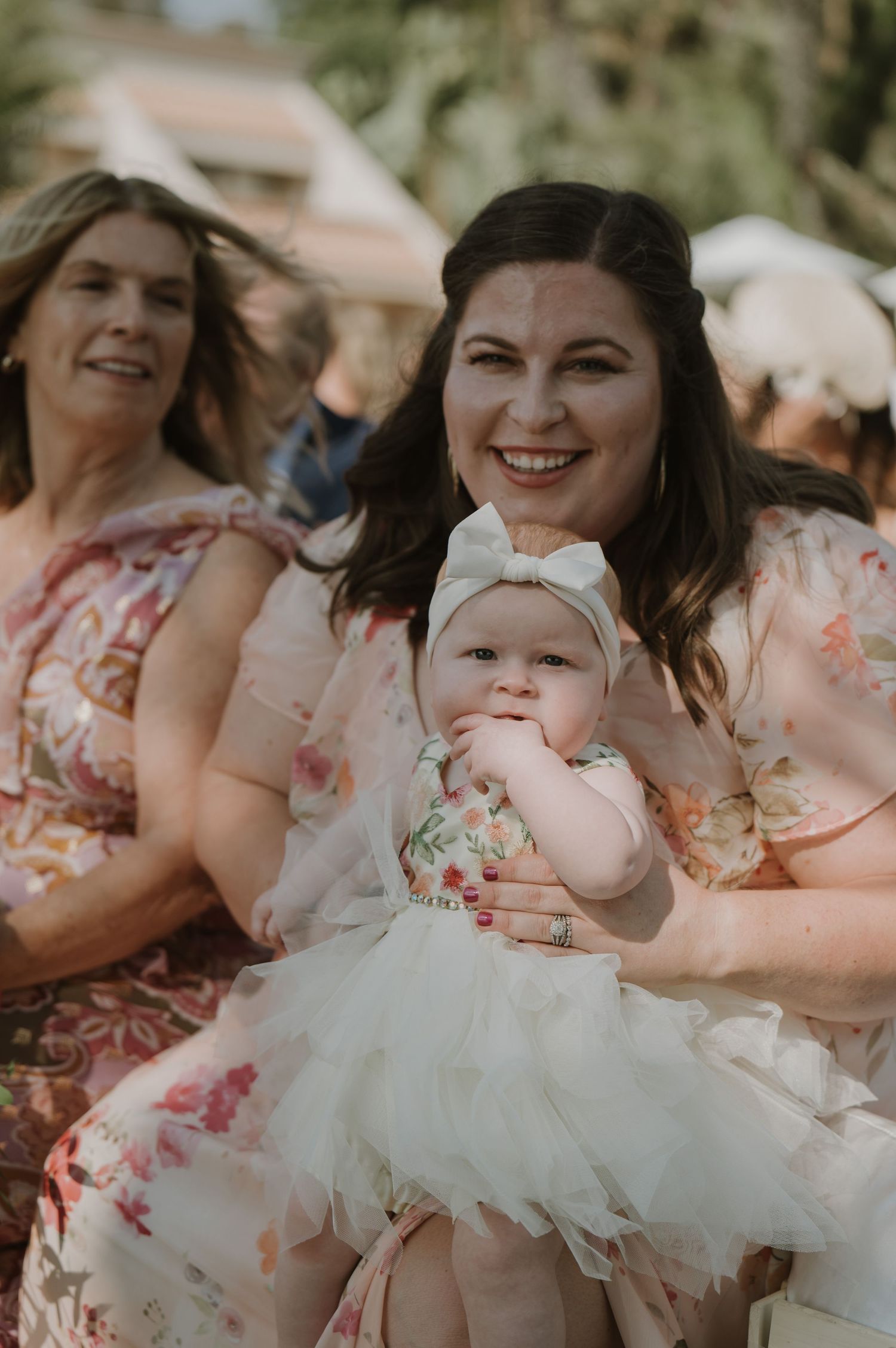 Wedding party members in floral pink dresses gather around a baby in a white dress.