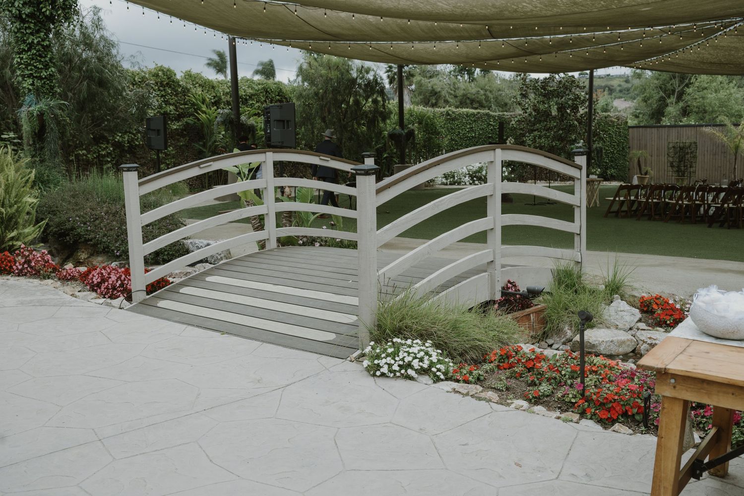 A decorative white wooden garden bridge surrounded by colorful flower beds in a landscaped outdoor patio area.