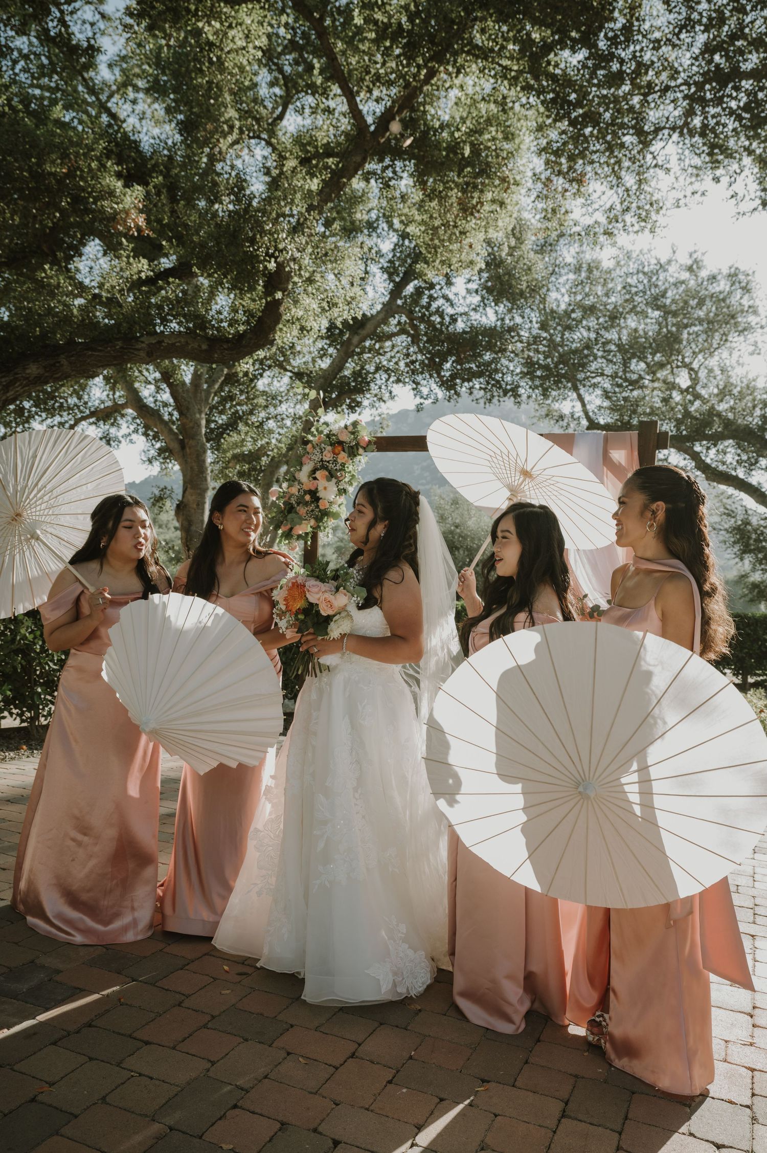 Bride and bridesmaids in blush pink gowns pose with white paper parasols under trees at outdoor wedding venue.