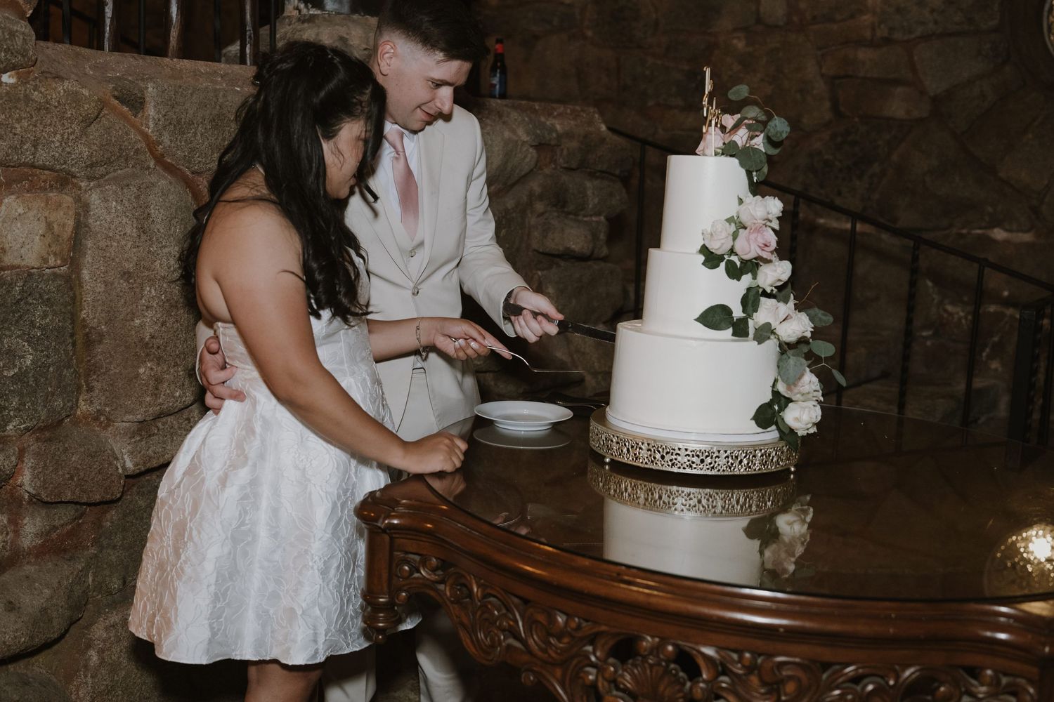 Bride and groom cutting a white four-tier wedding cake decorated with pink roses in a stone venue.