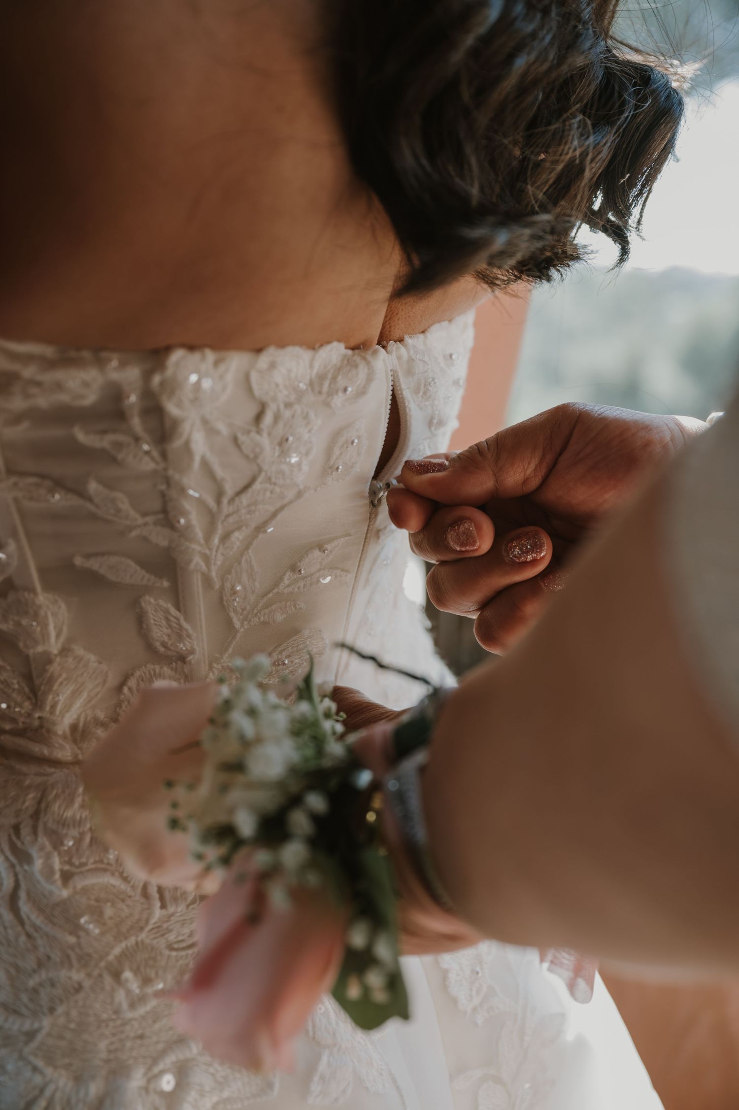 Close-up of bridesmaid fastening buttons on back of intricate lace bridal gown, wearing floral wrist corsage.
