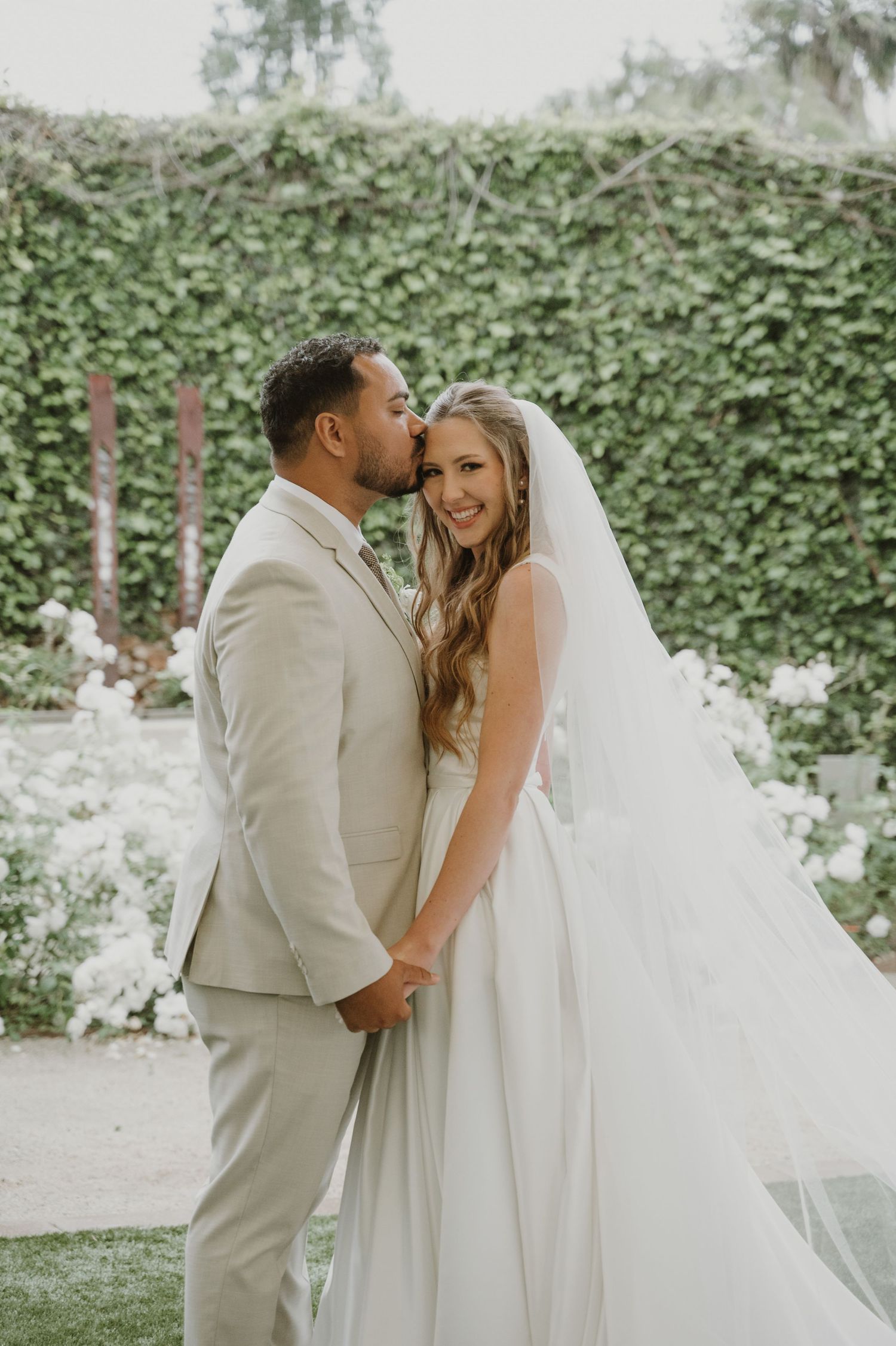 Groom in beige suit kisses bride's cheek as she smiles in white wedding gown and veil in garden.