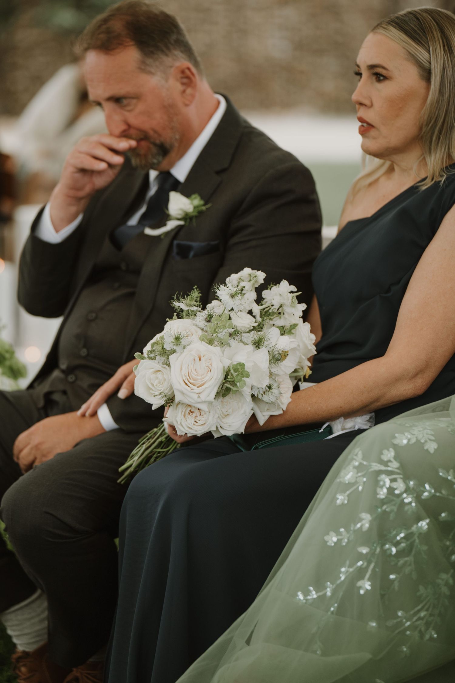 Emotional wedding guest in dark suit holding white rose bouquet during outdoor ceremony.
