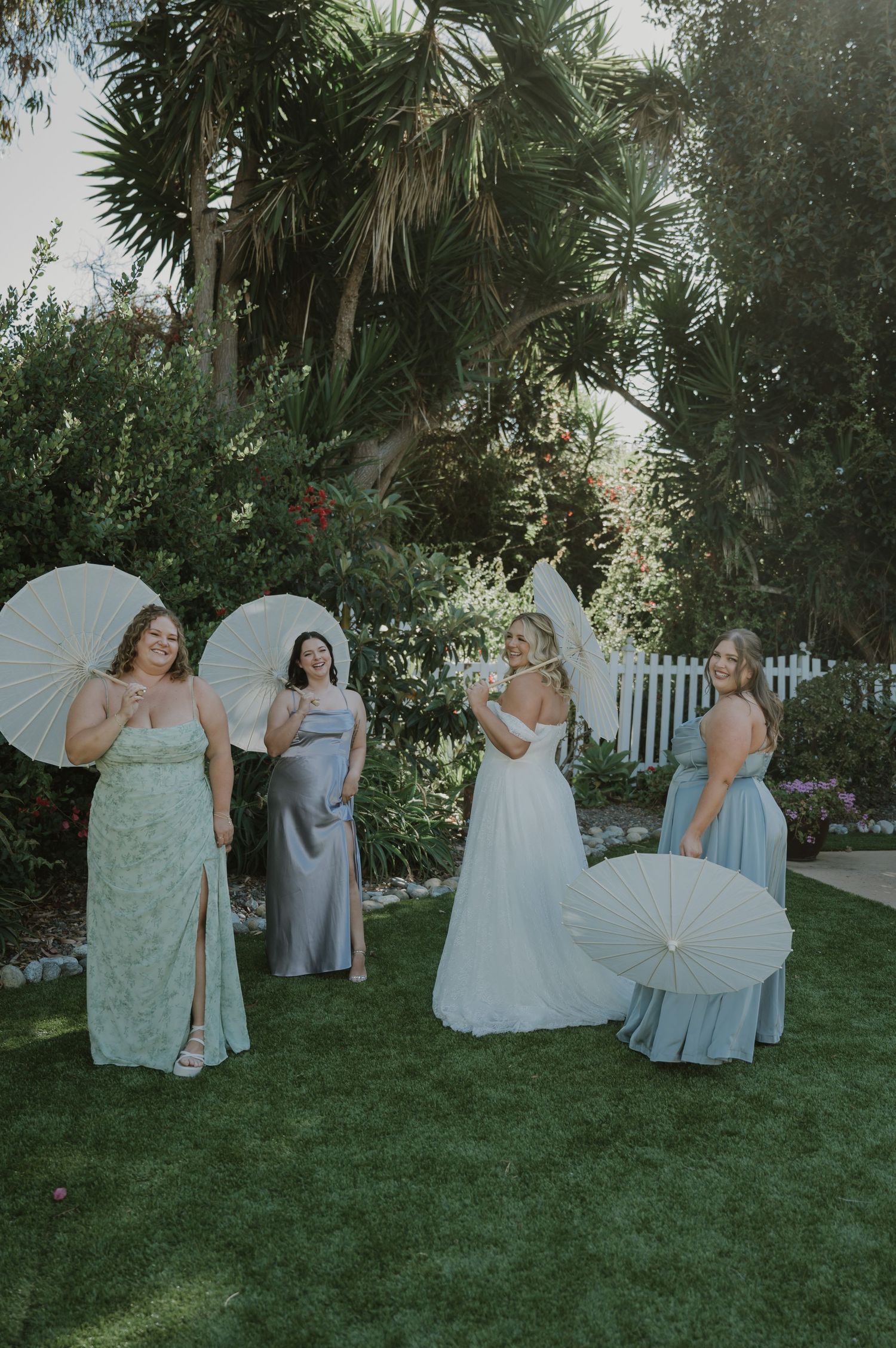 Bride in white gown and bridesmaids in sage and silver dresses holding parasols in a garden.