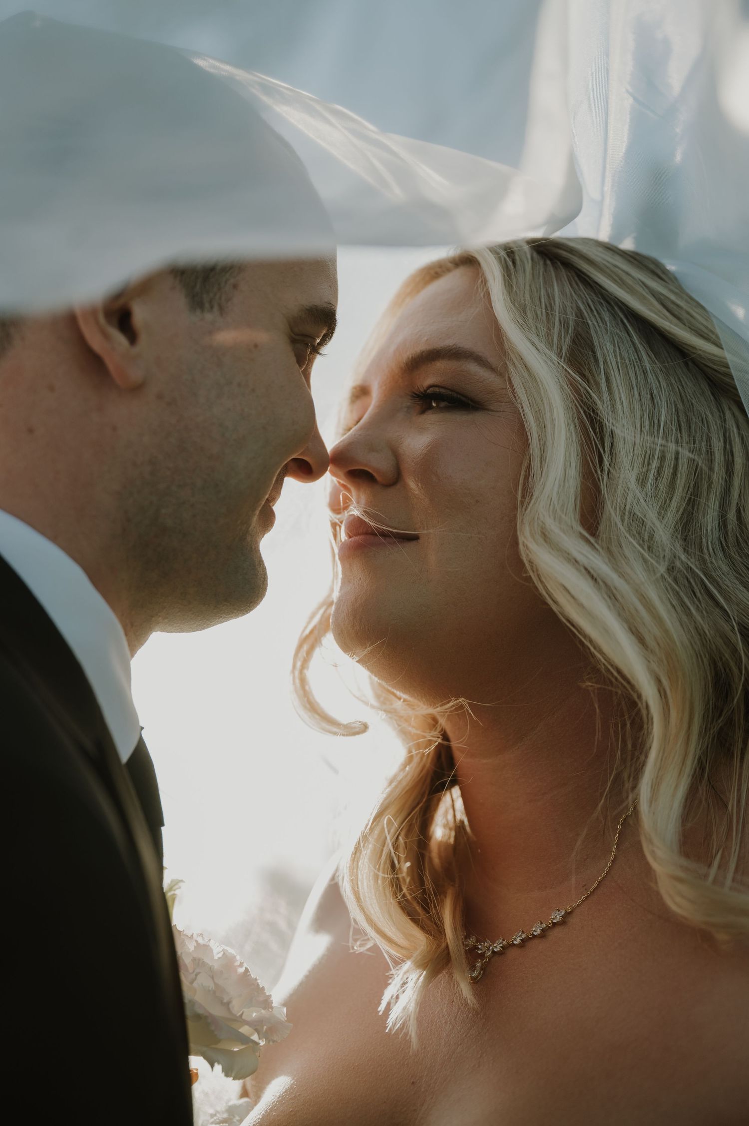Bride and groom sharing an intimate moment under a veil, bathed in golden backlight on their wedding day.