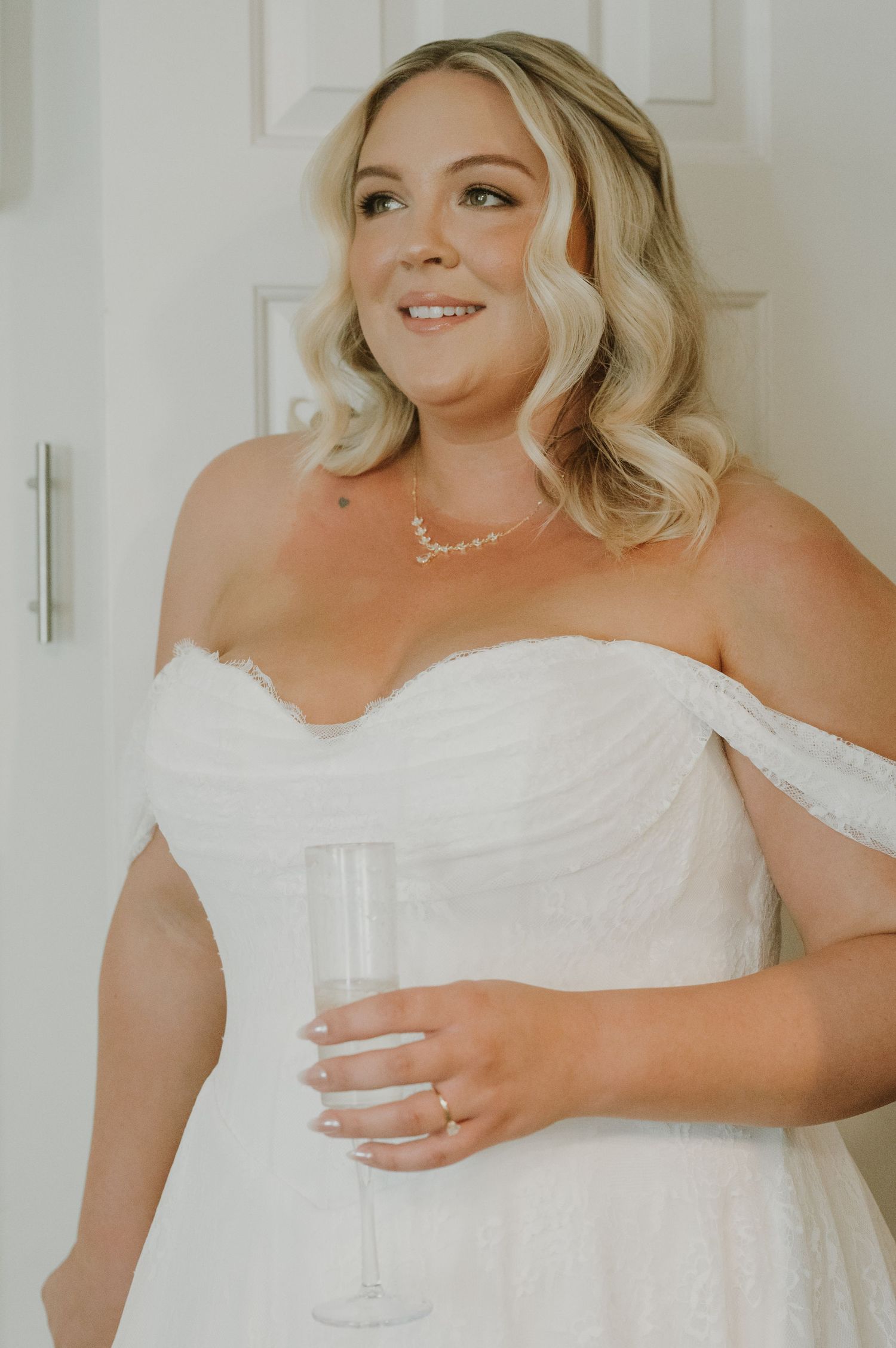 Beautiful blonde bride in off-shoulder lace wedding dress holding champagne flute, wearing pearl necklace and engagement ring.