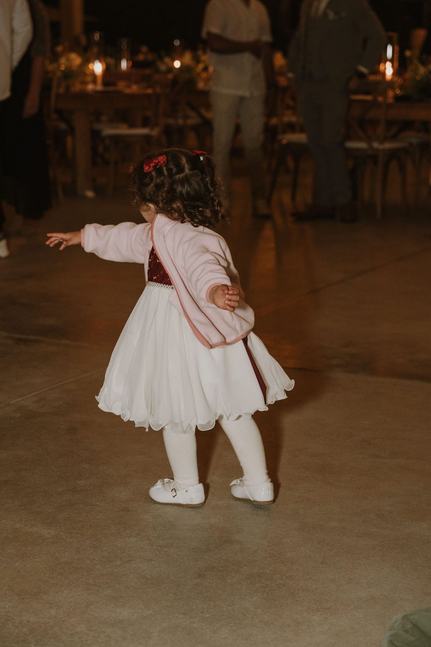 Young flower girl in white dress and pink jacket dancing at an outdoor wedding reception at night.