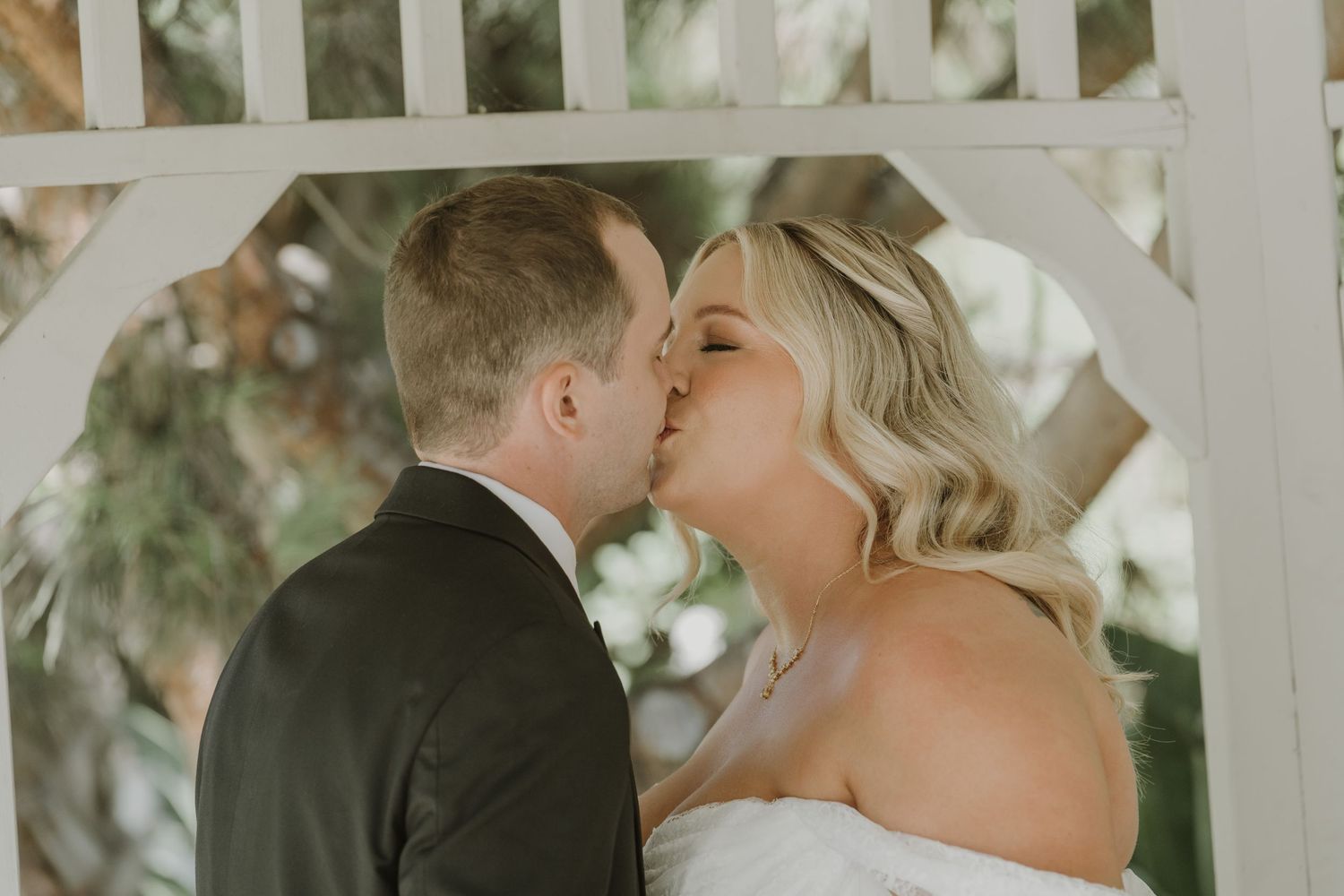 A bride and groom share a romantic kiss under a white wooden gazebo on their wedding day.
