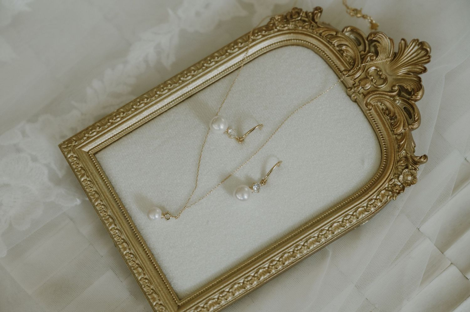 Gold ornate frame displaying delicate pearl necklace and earring jewelry set on white fabric background.