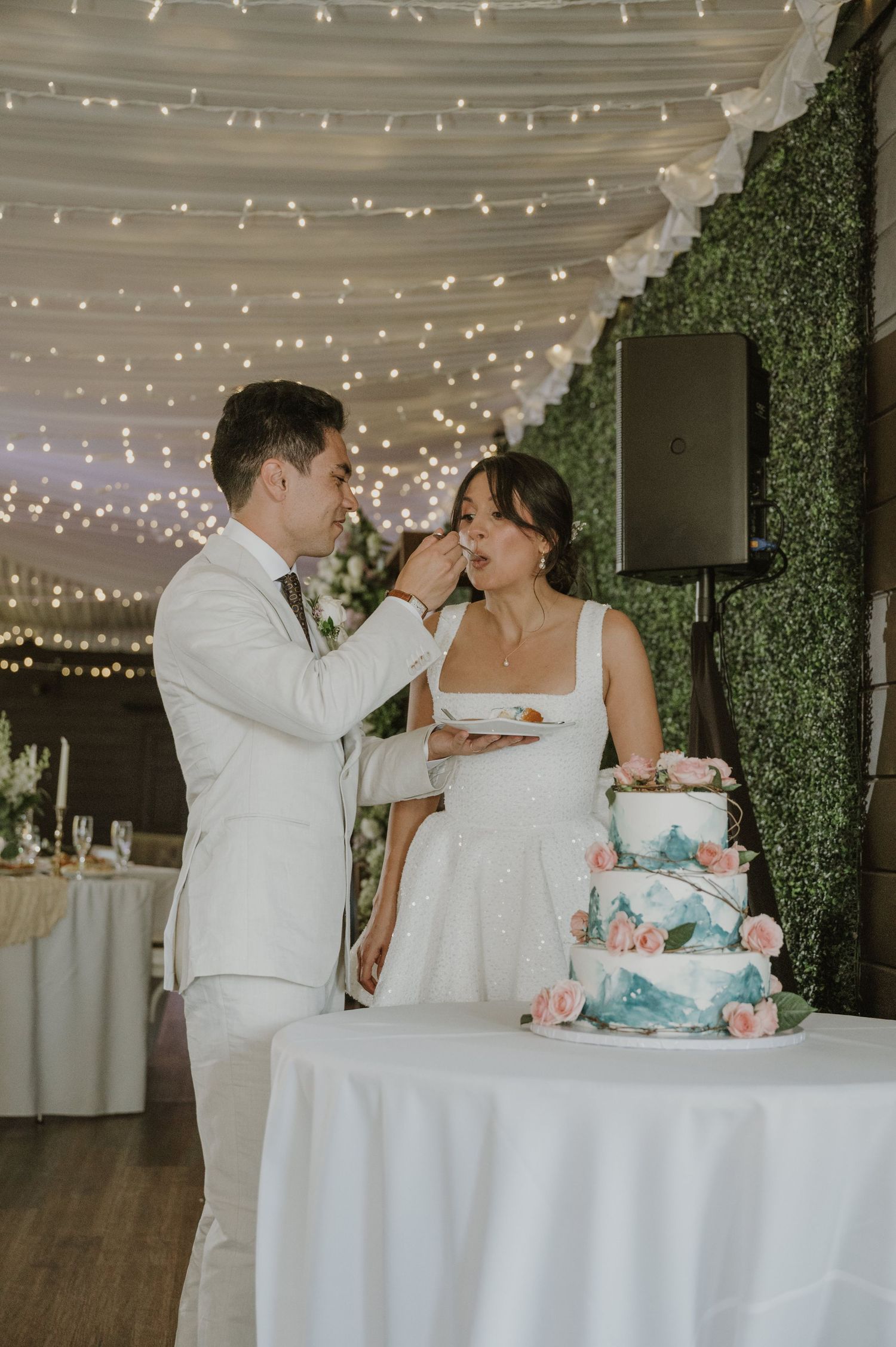 Groom in white suit feeds wedding cake to bride under fairy lights beside tiered blue floral cake.