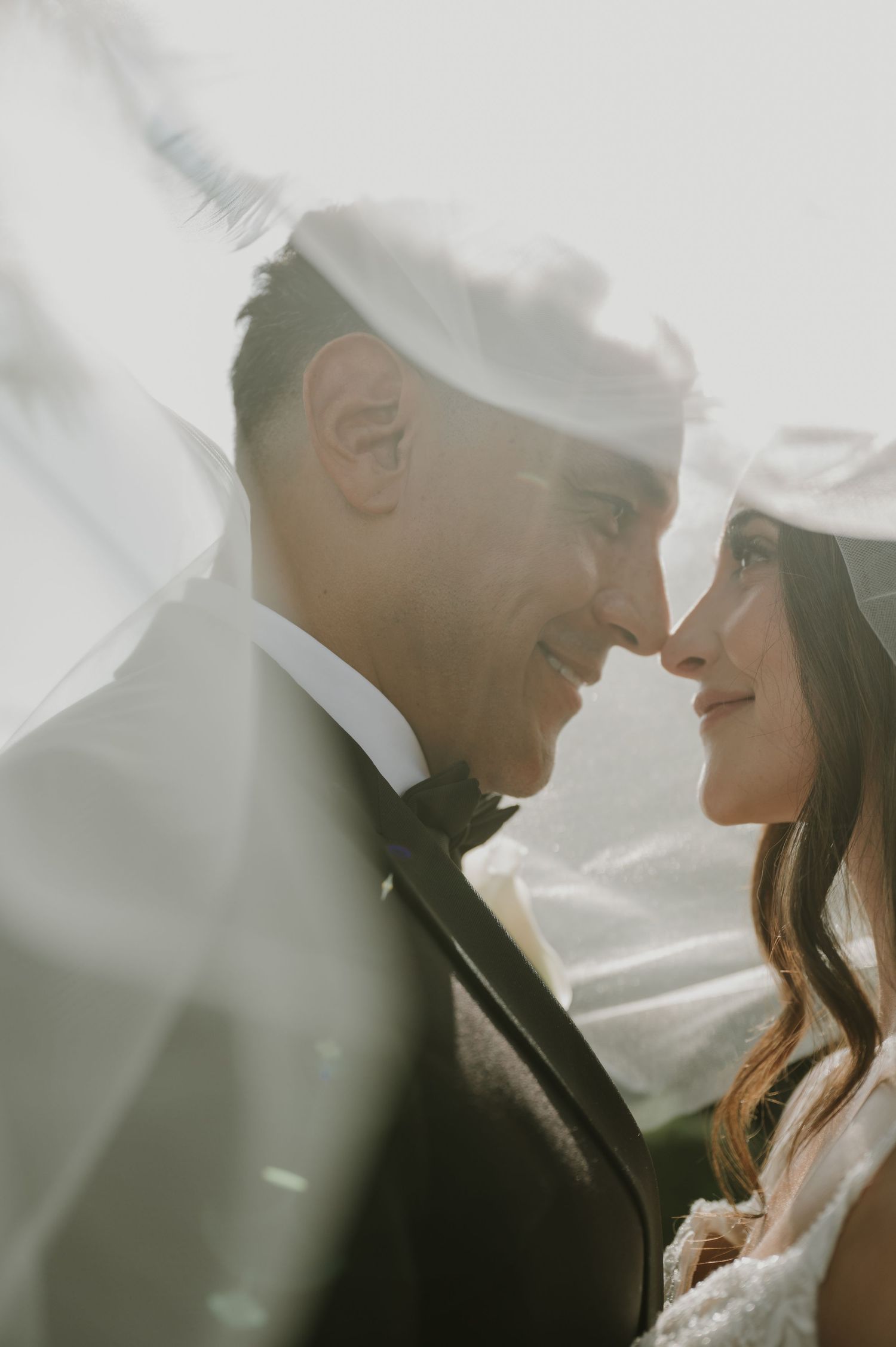 Bride and groom sharing an intimate moment under a flowing veil, smiling at each other on their wedding day.