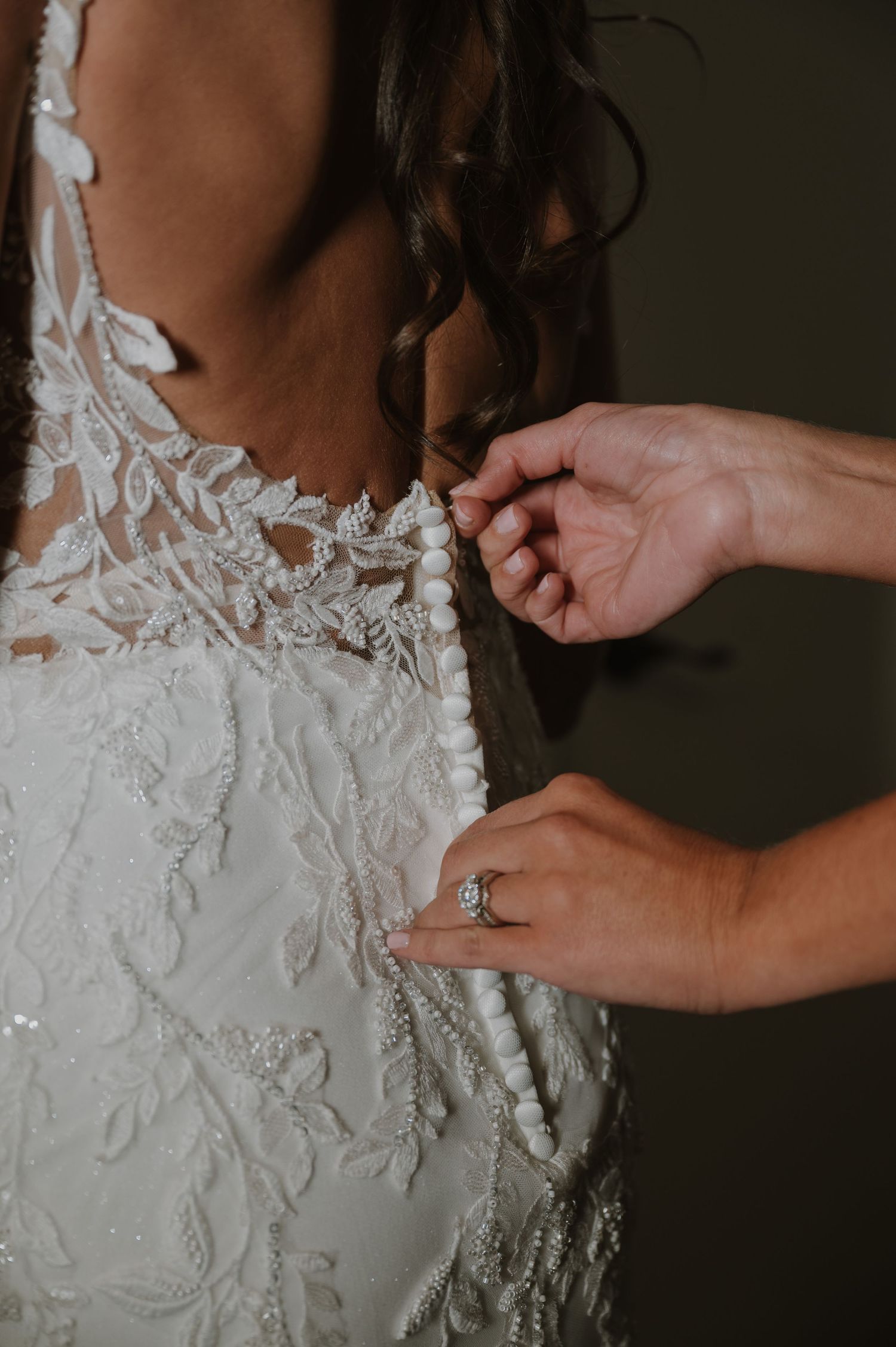 A bride in a beaded lace wedding gown has her back buttons fastened by a helper wearing a diamond ring.