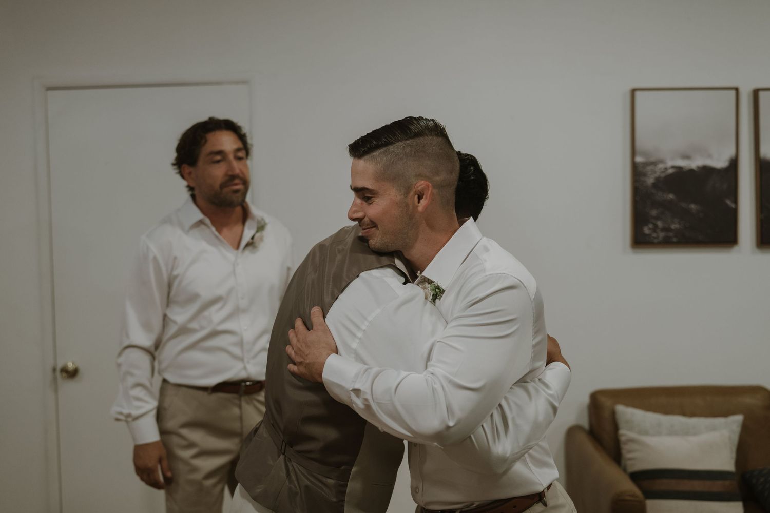 Groomsmen sharing a warm hug while another man watches during pre-wedding preparations indoors.