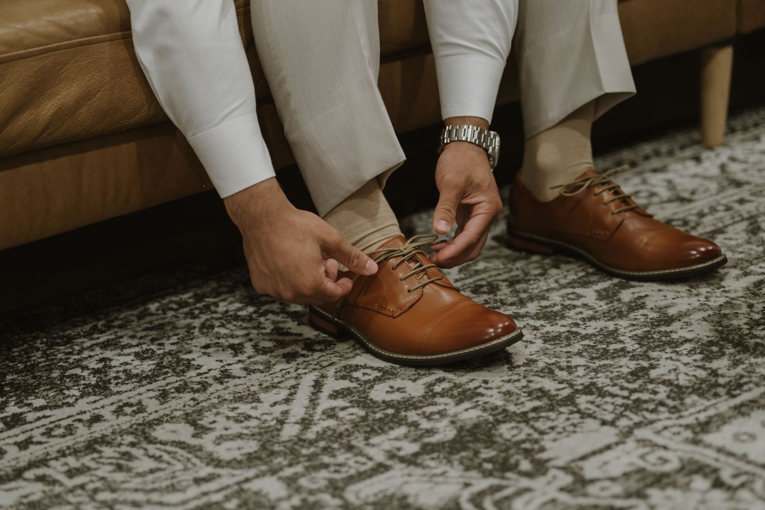 Man in white trousers tying brown leather Oxford dress shoes while seated, wearing a silver watch.