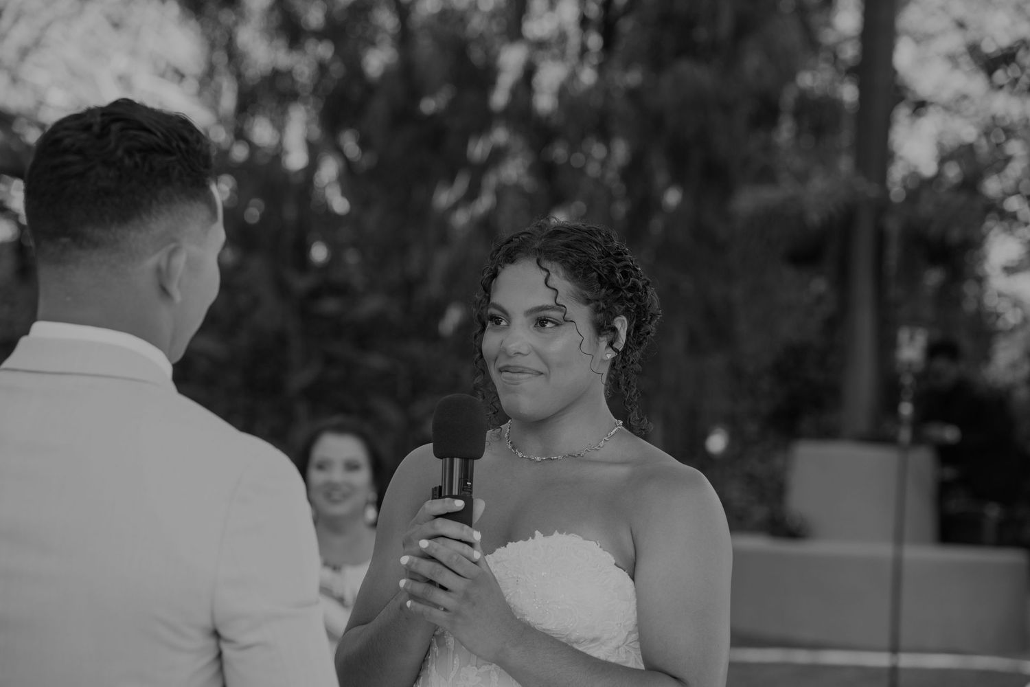 Black and white photo of a bride smiling while reciting wedding vows into a microphone at an outdoor ceremony.