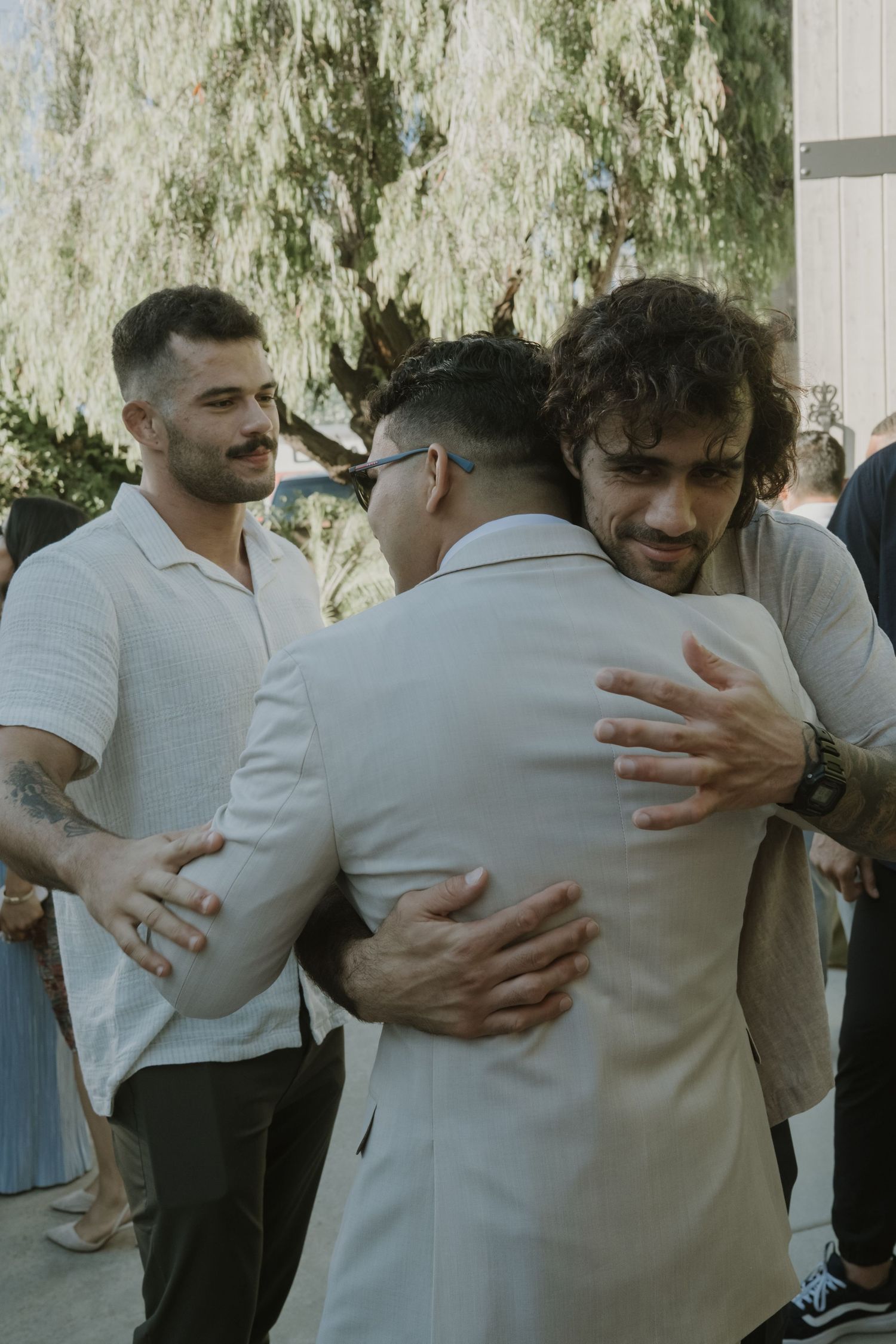 Two men share an emotional hug at an outdoor wedding while a smiling guest with tattoos looks on.