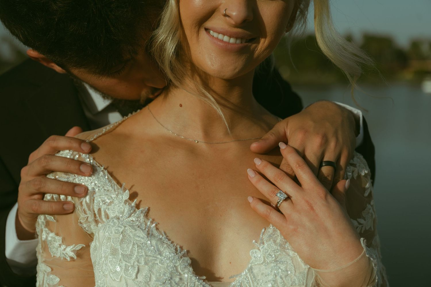 Bride and groom embracing, showcasing wedding rings and lace bridal gown near a scenic waterfront.