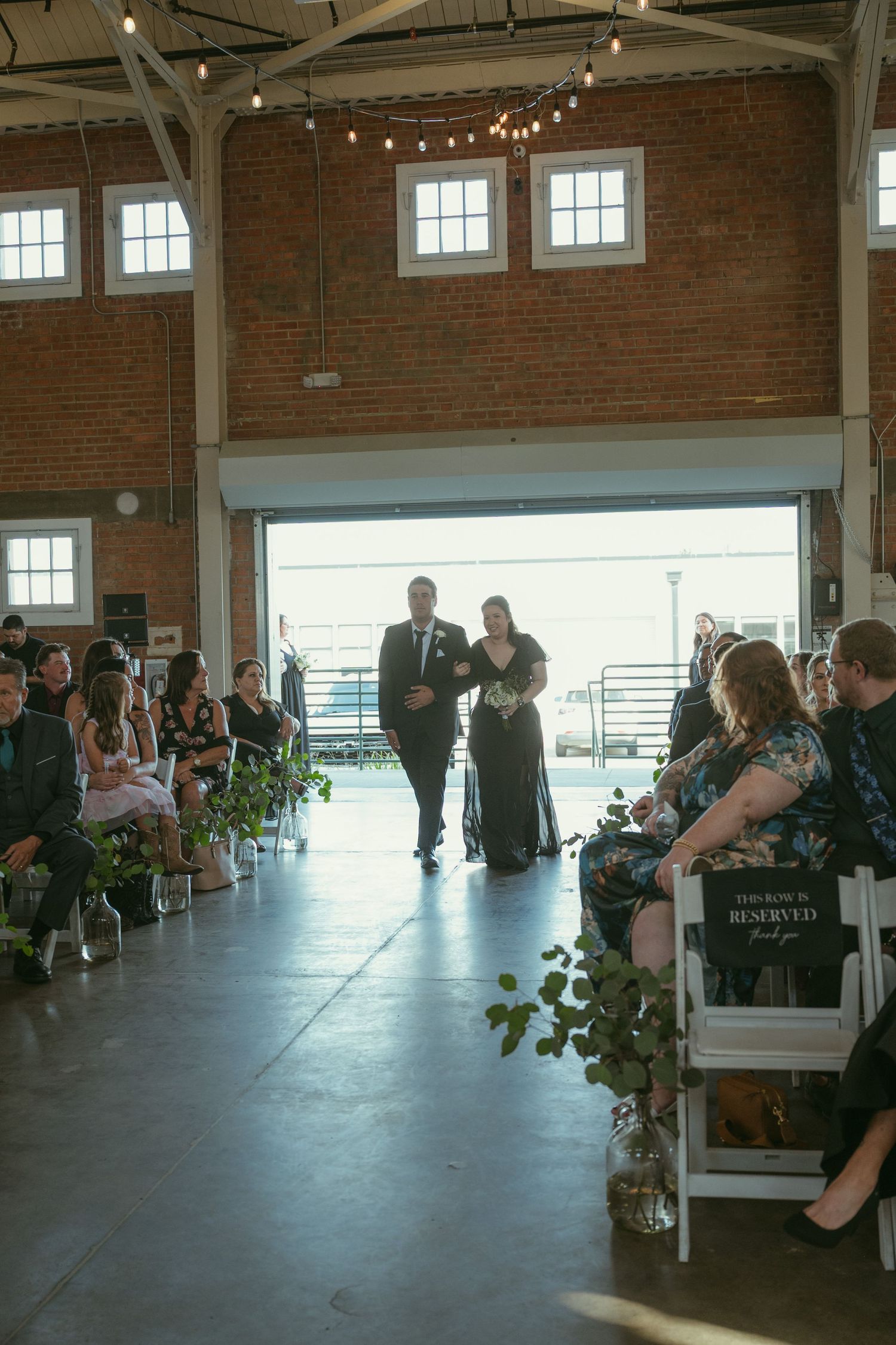 Couple walks down the aisle at an industrial brick wedding venue with string lights and seated guests.