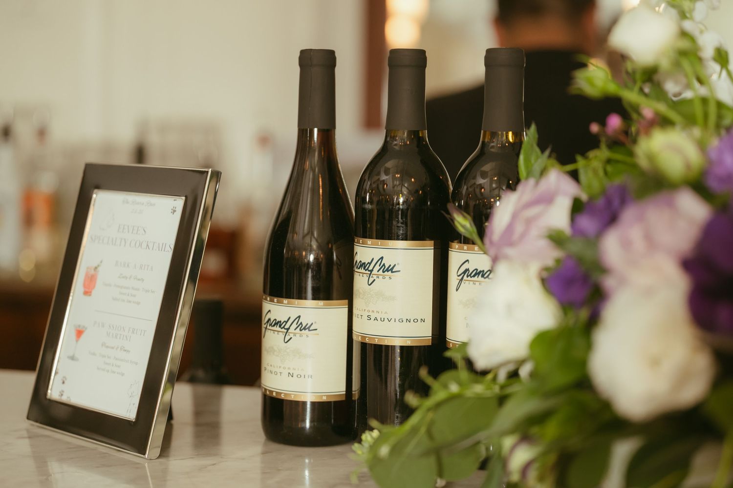 Three Grand Pre wine bottles displayed beside a purple and white floral arrangement at a wedding.
